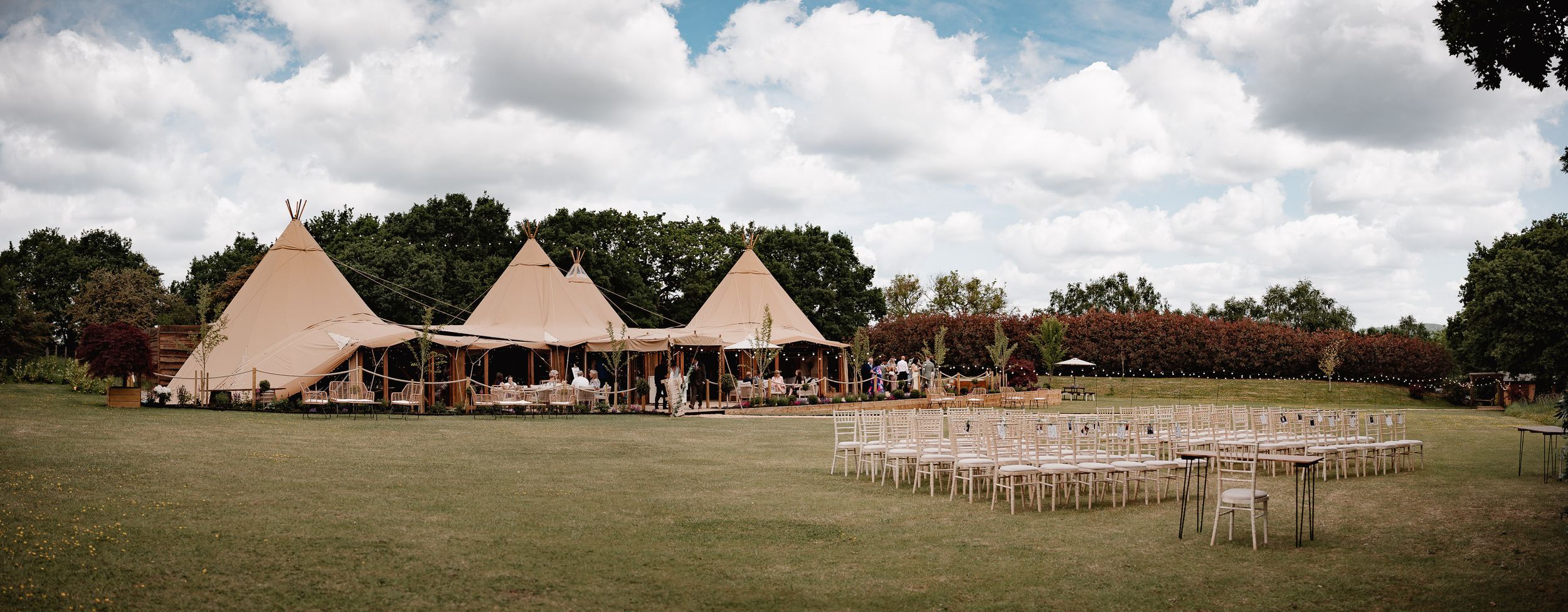Panoramic view of tipi-style wedding tents set up in a grassy field with white chairs arranged for an outdoor ceremony.