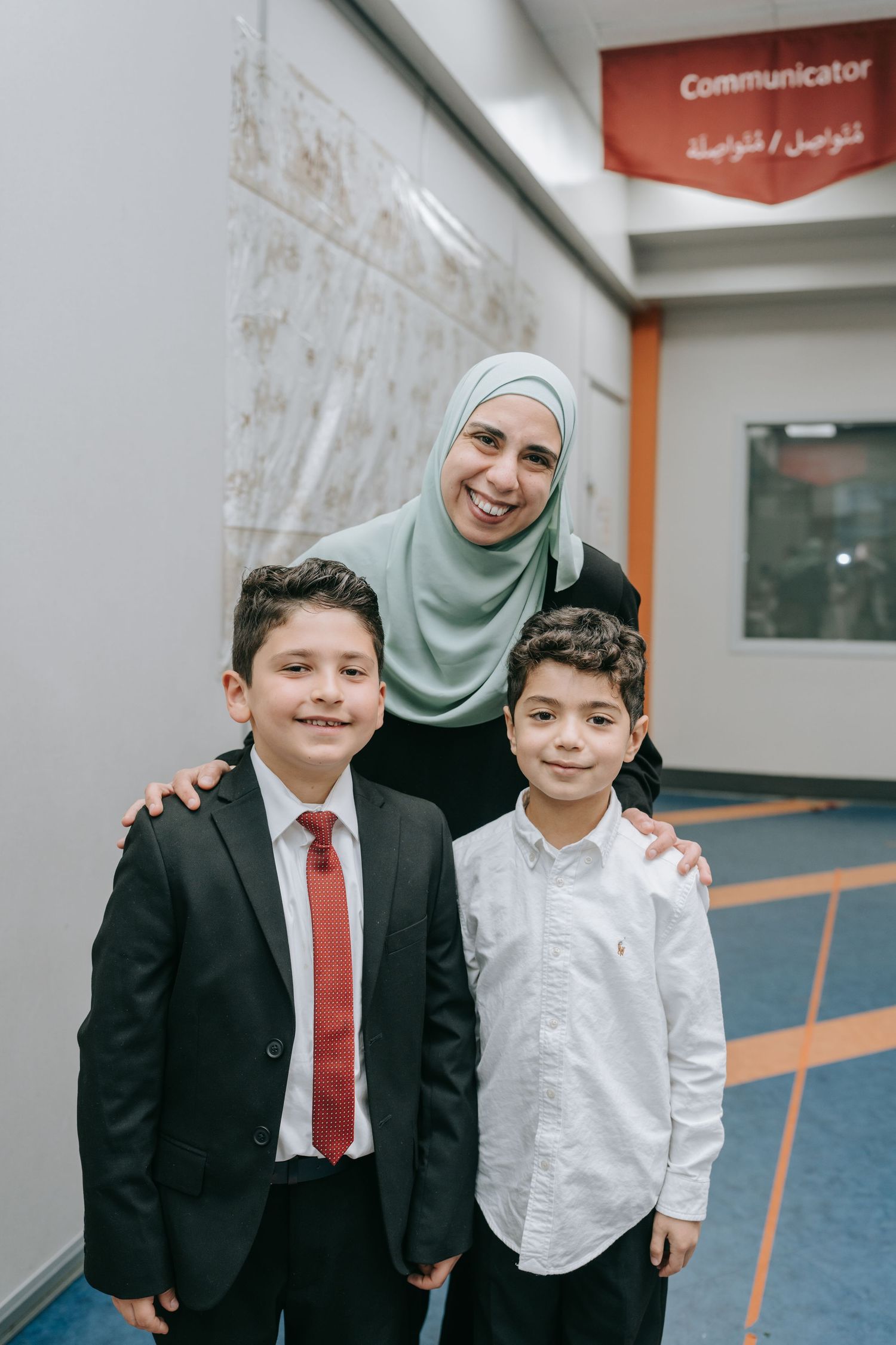 A person wearing a mint green hijab stands warmly with two young boys in a hallway near a sign.