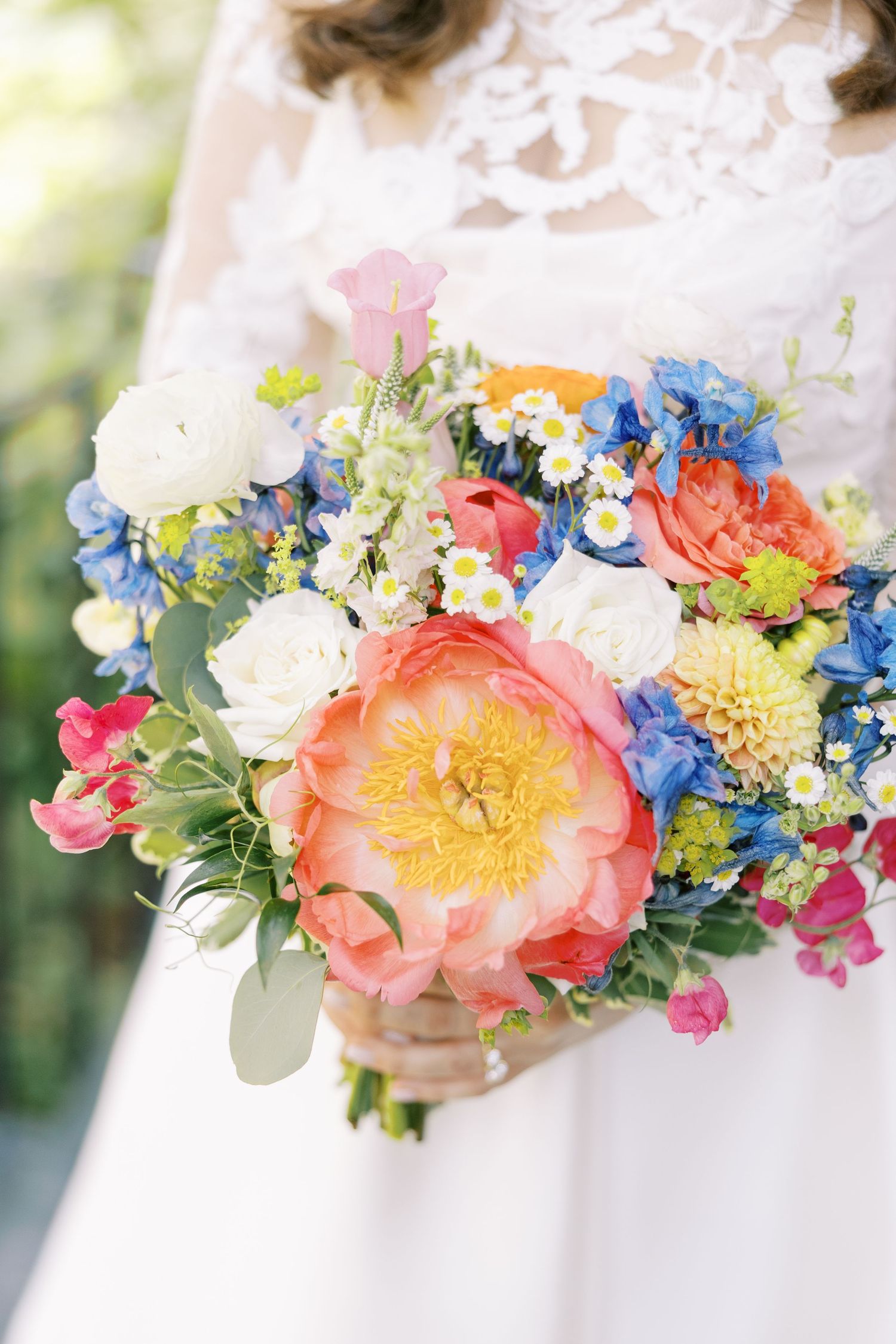 A vibrant bridal bouquet featuring coral peonies, blue delphinium, white roses and yellow accents against a white lace background.