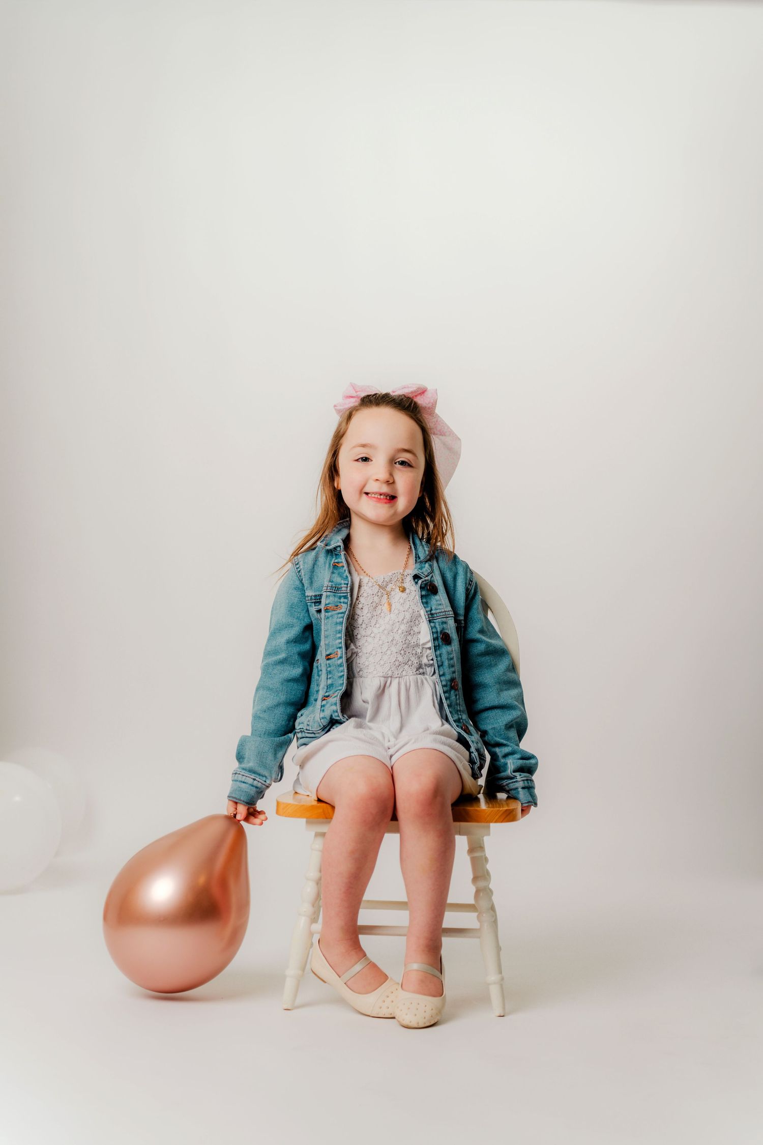 A child in light blue overalls stands barefoot next to a white chair with a birthday cake displaying the number 2.