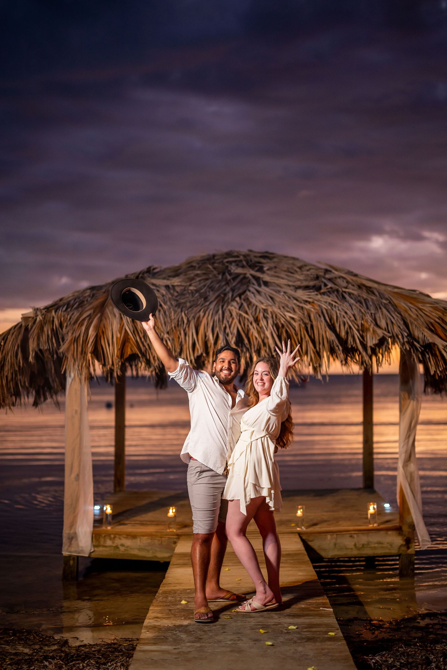 Two people celebrate joyfully under a thatched palapa on a wooden pier at sunset with glowing candle lights.