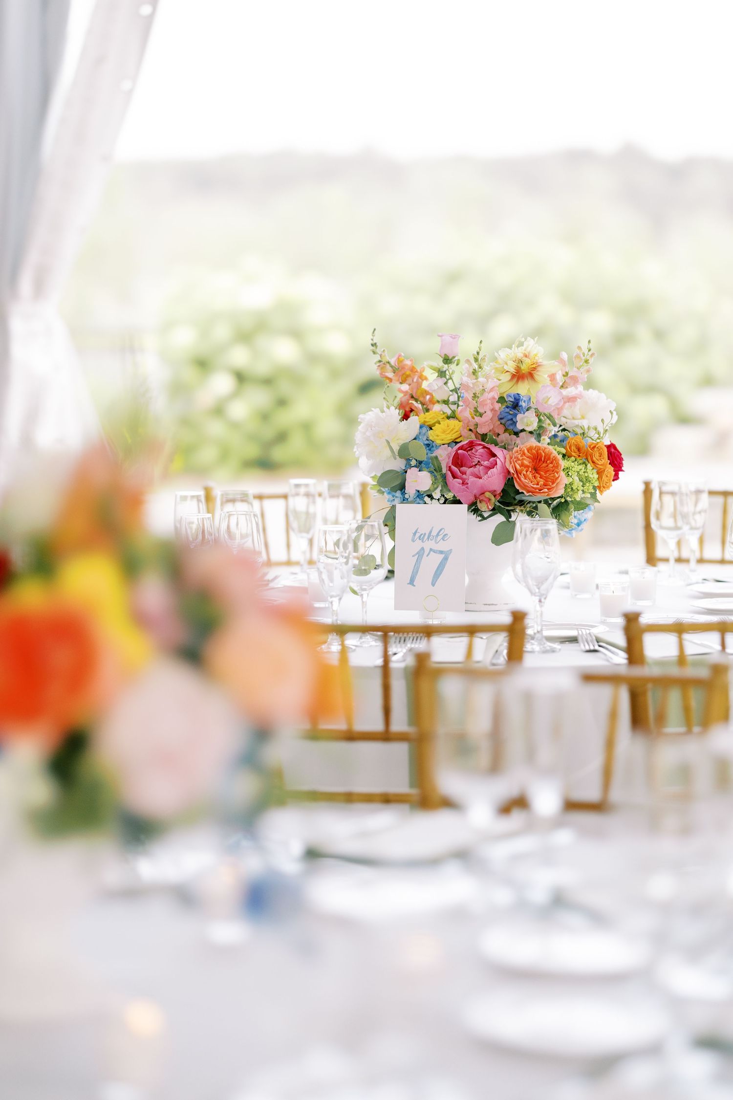Elegant wedding reception table setting with colorful floral centerpiece, gold chairs and table number under white tent.