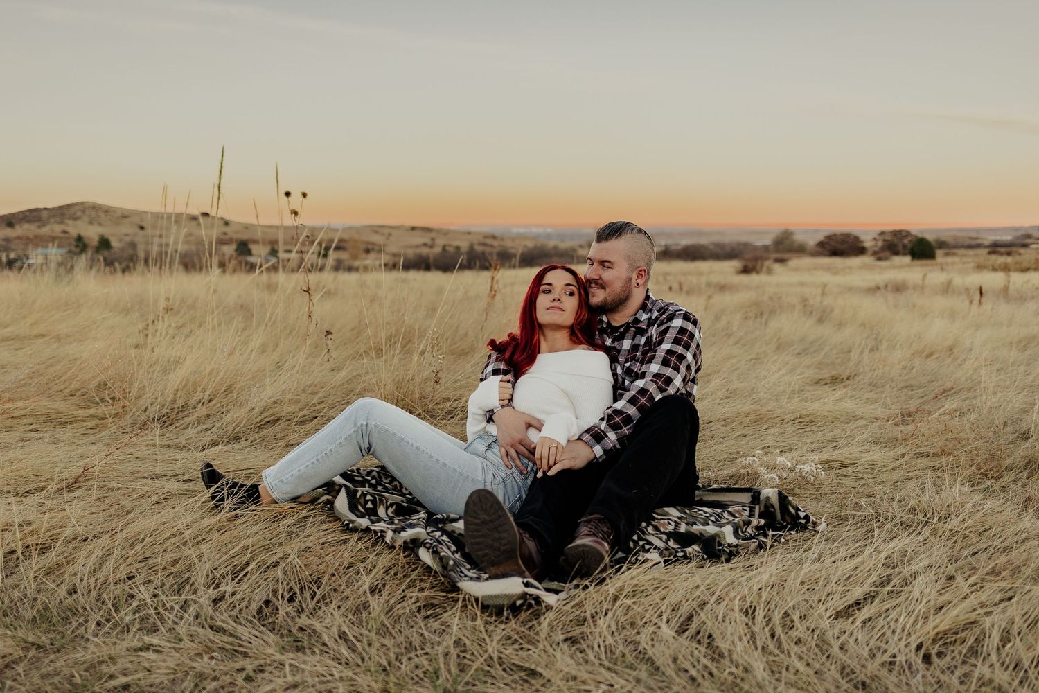 A couple rests in a golden prairie field during sunset casting long shadows across the dried grass.