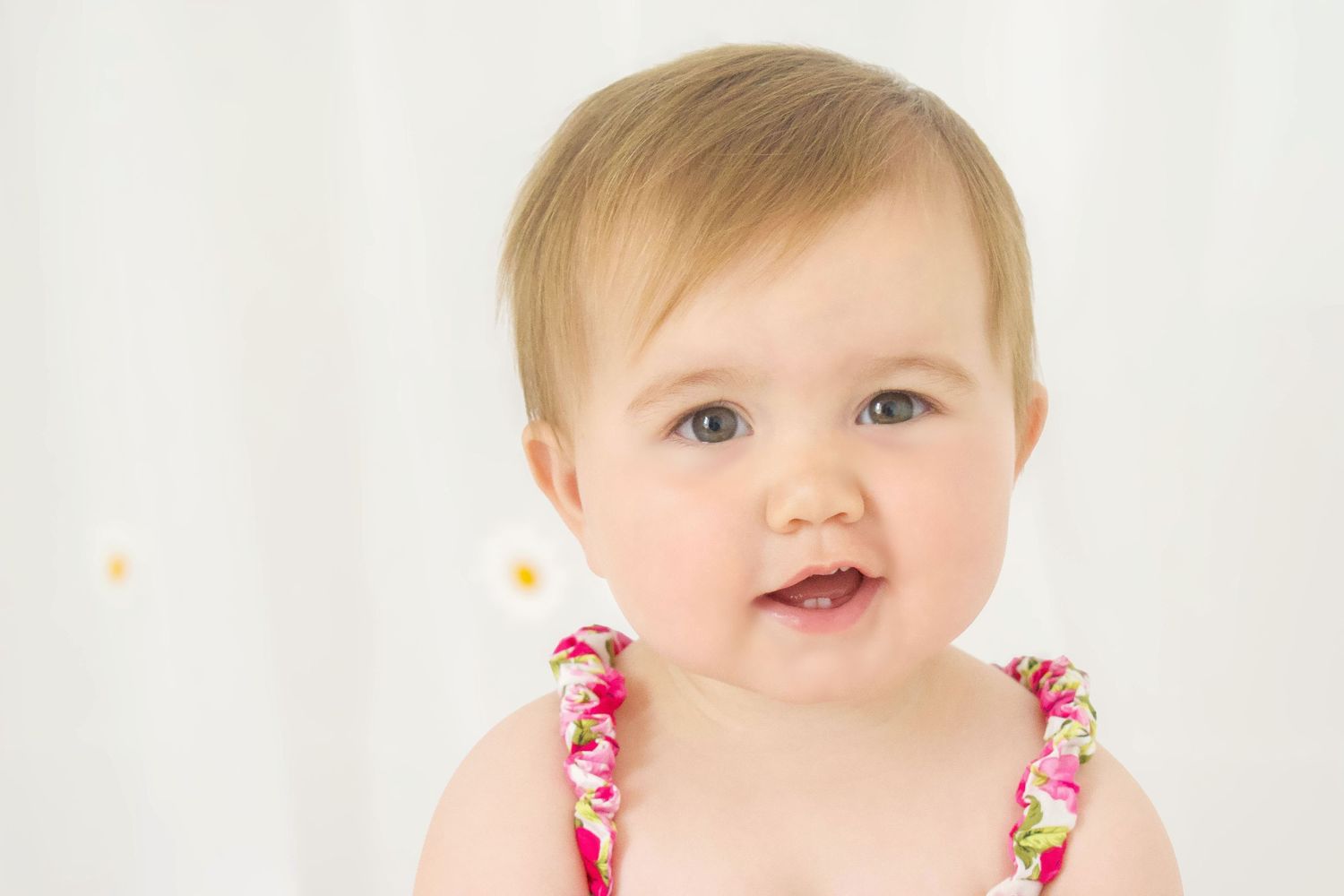 Baby in pink floral dress smiling at camera against white background.