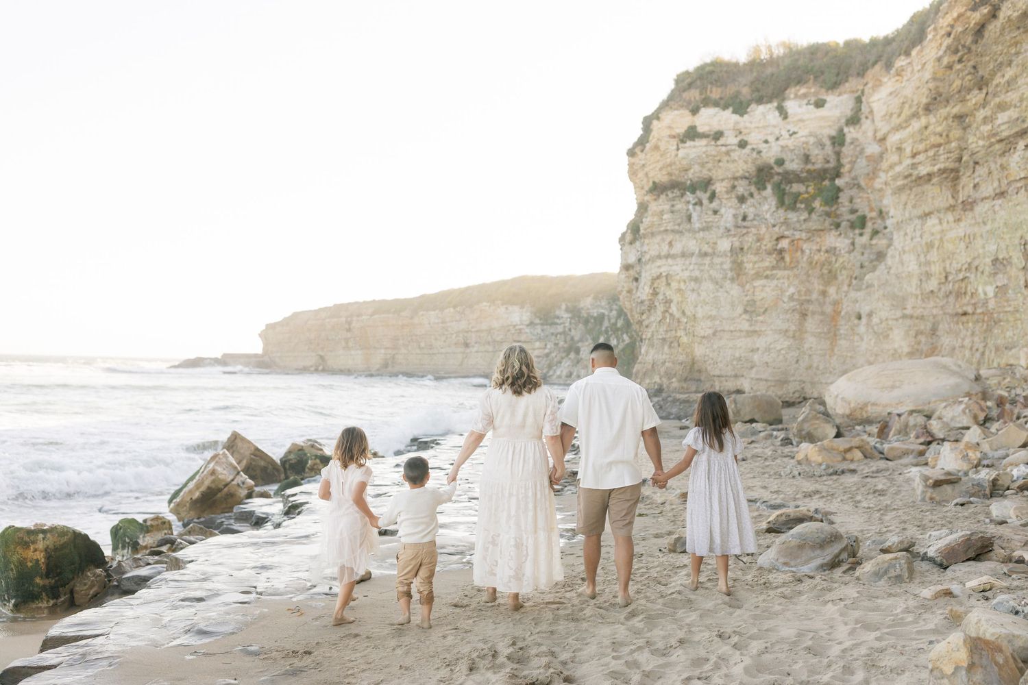 Family holding hands while looking at ocean waves crashing against rocky cliffs on a sunny beach captured by Chelsee Rawe Photography.