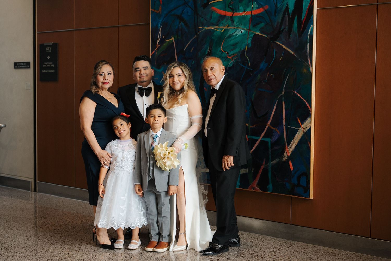 A wedding party in formal attire poses together in front of a modern art piece in an indoor venue.