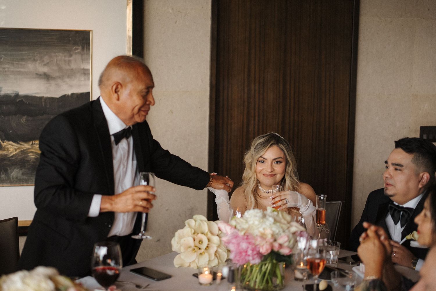 Wedding reception guests share a lighthearted moment at an elegant table with floral centerpieces and formal attire.