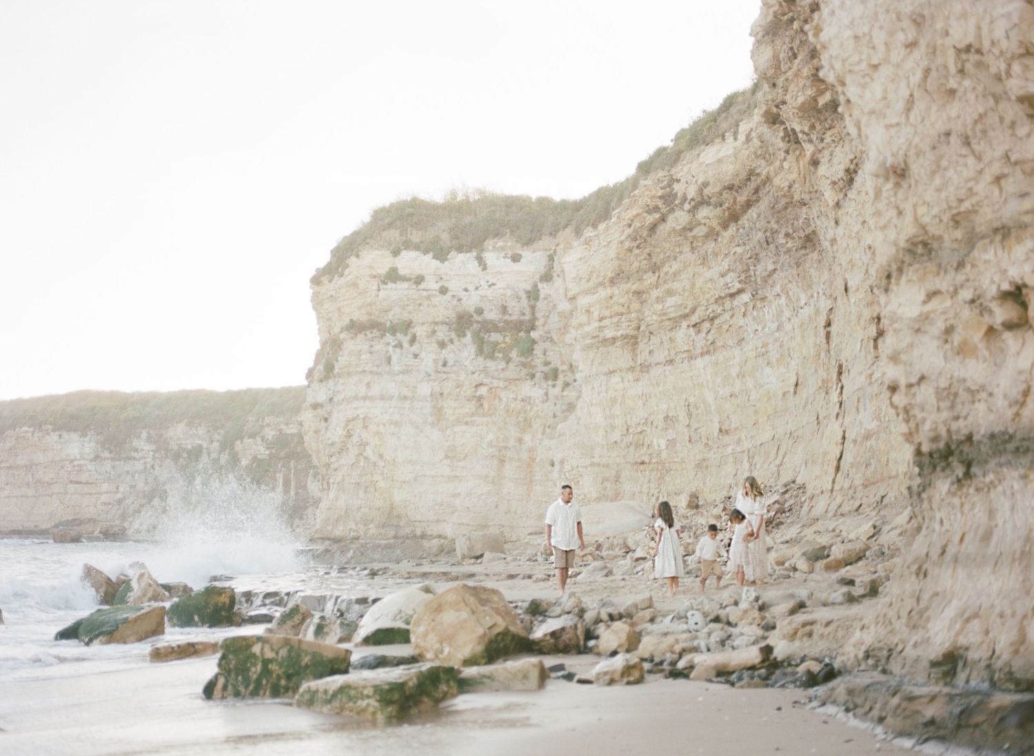 A figure stands near the shore beneath towering white chalk cliffs with waves crashing on rocky beach film by Chelsee Rawe .