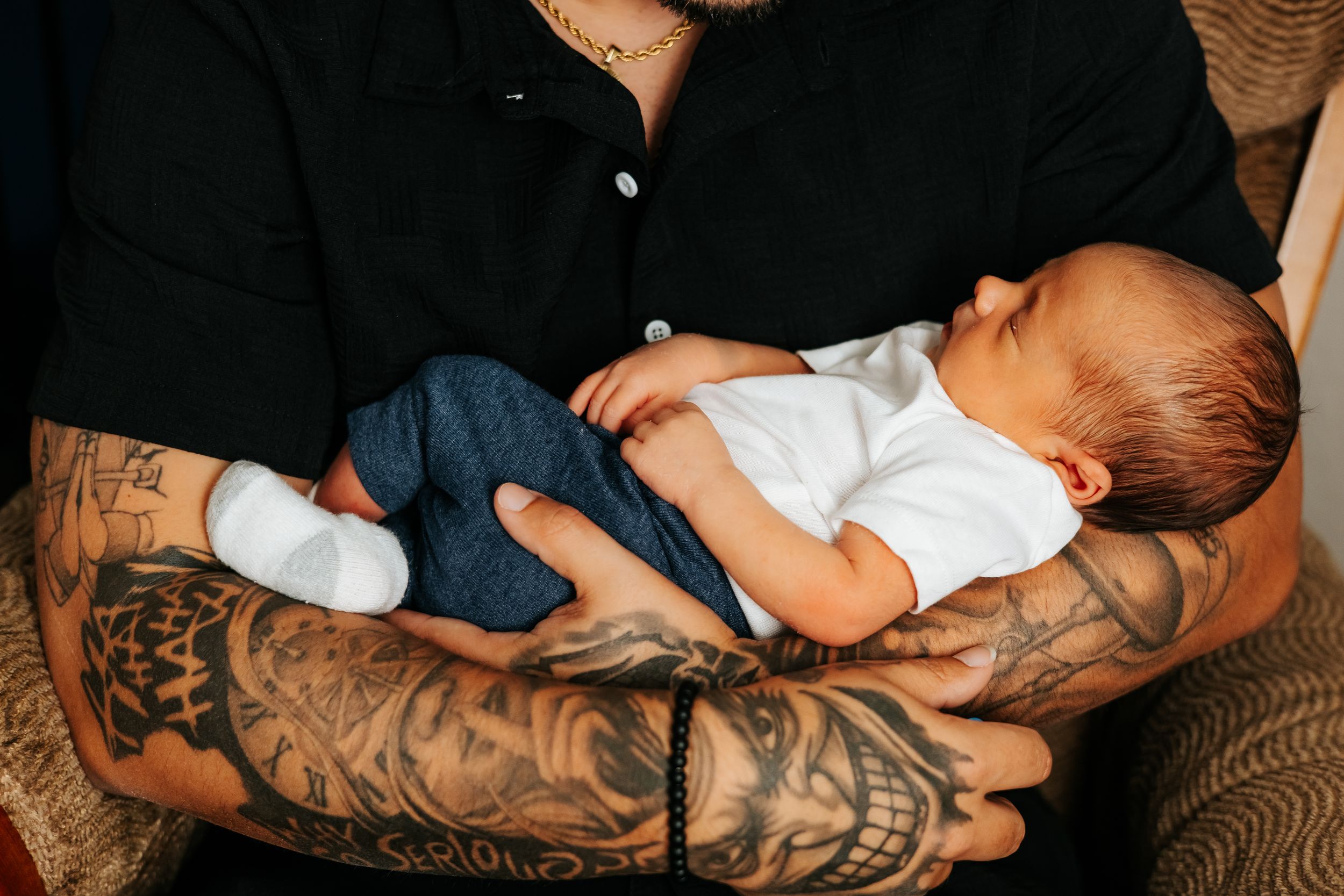 Tattooed arms hold sleeping newborn baby wearing white onesie.