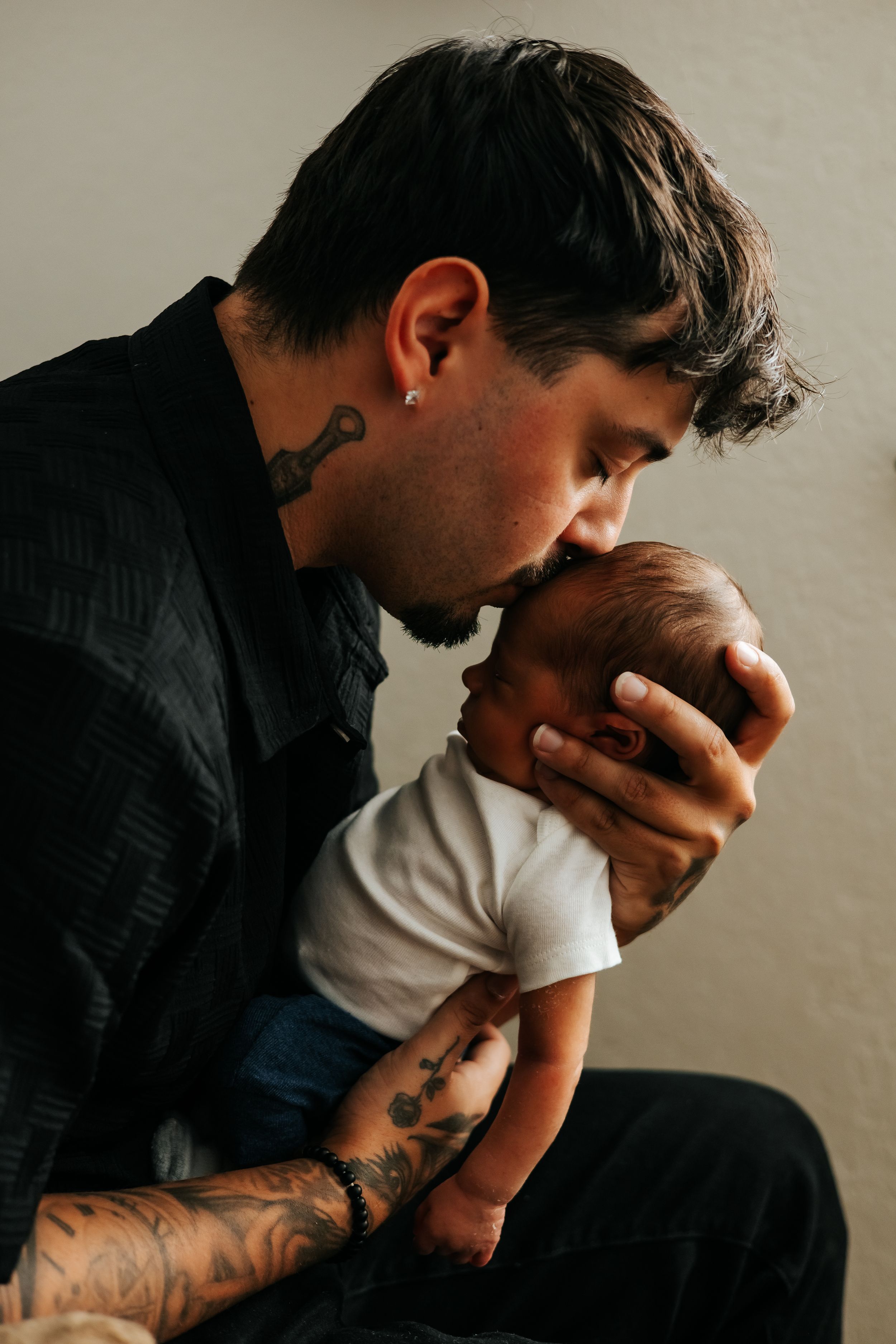 Parent with tattoos gently kisses newborn baby's head in intimate portrait.