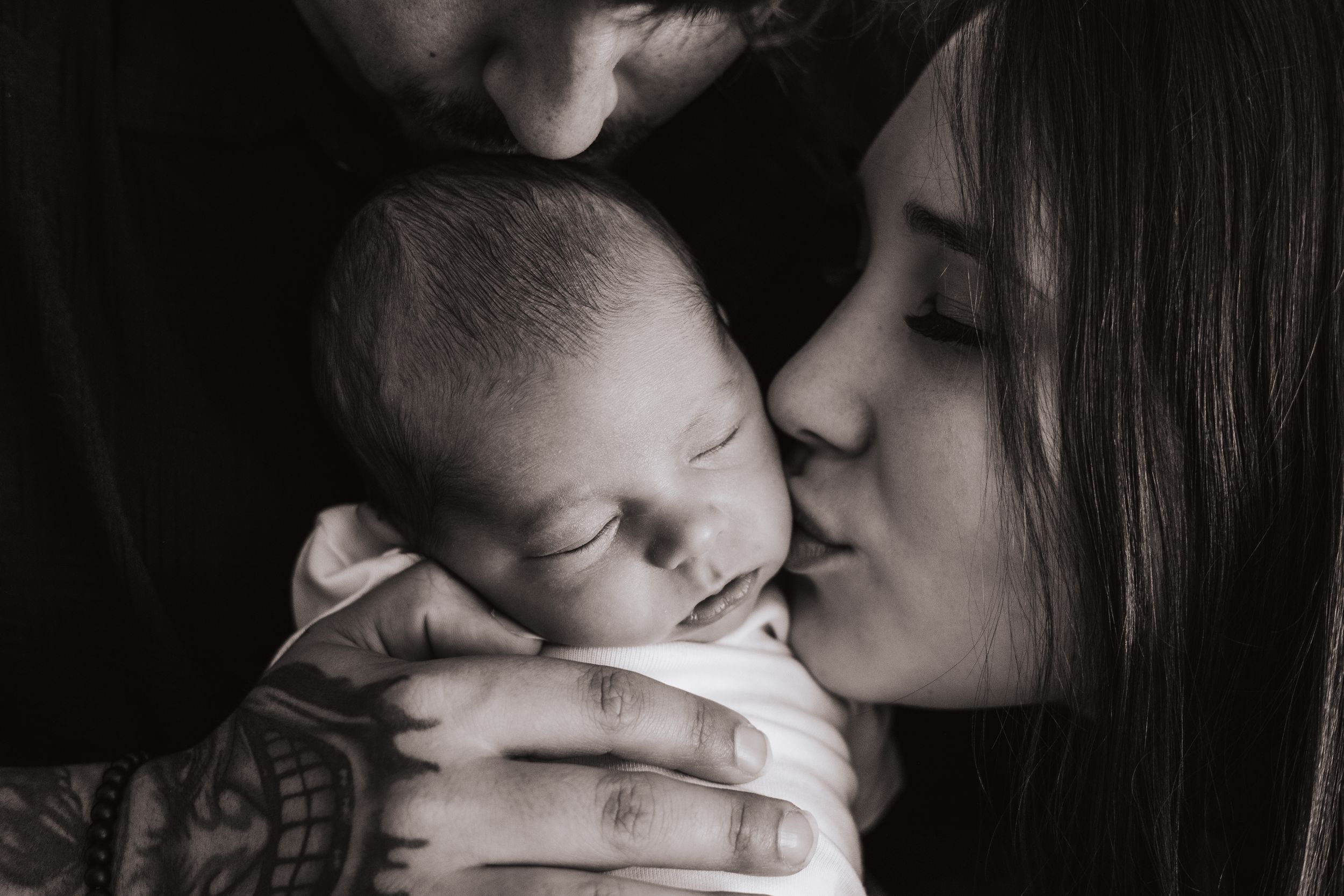 An intimate black and white portrait of a mother kissing her sleeping newborn.