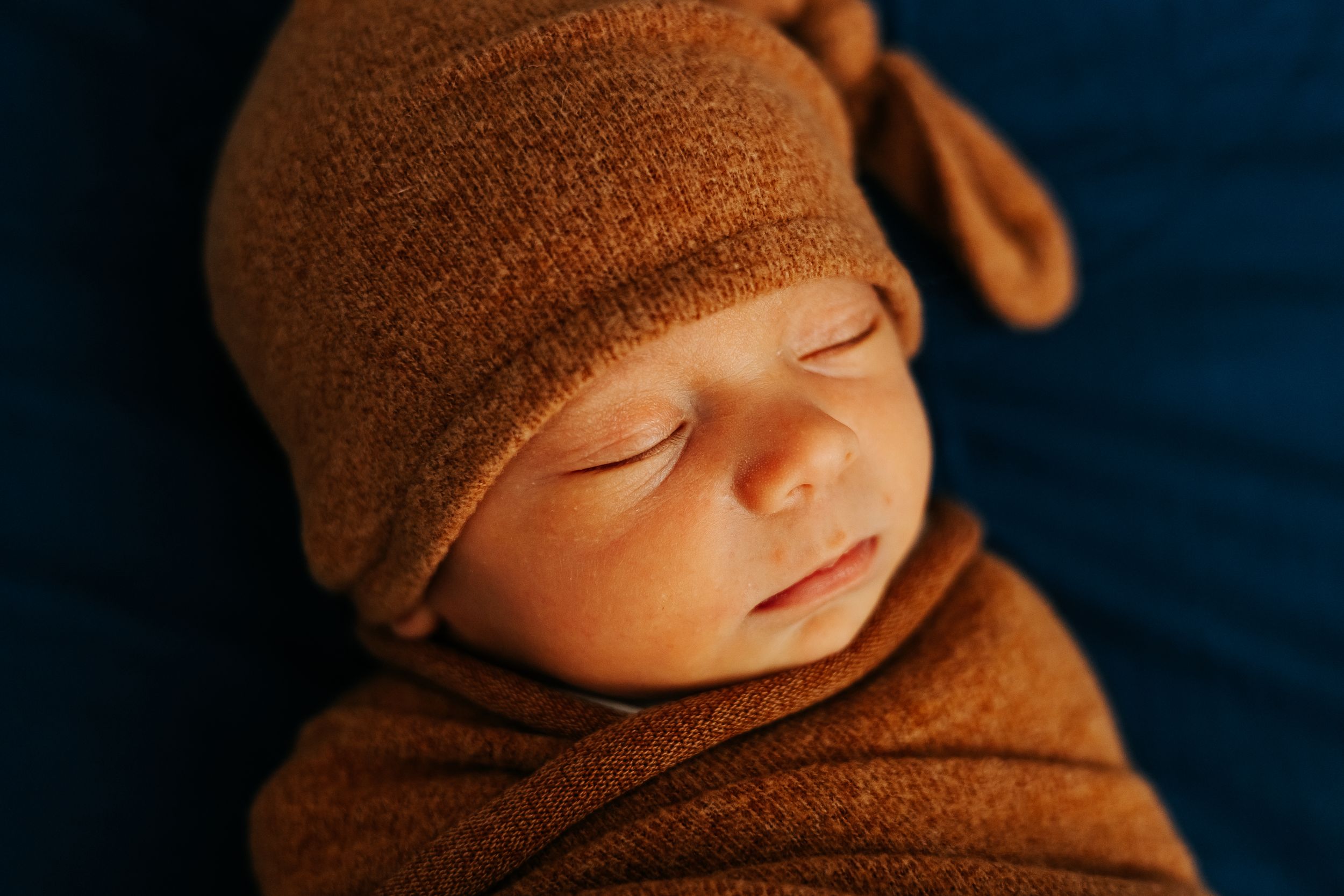 Sleeping newborn wearing a cozy brown knit hat in warm lighting.
