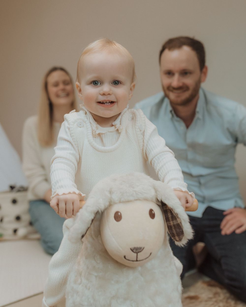 Smiling baby holding a plush lamb toy while sitting with family on a light colored background.