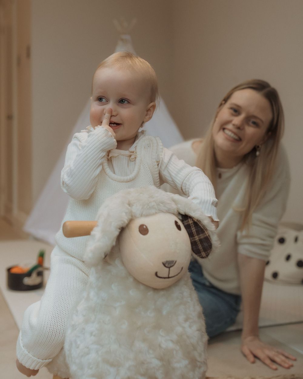 A toddler in white sits on a plush sheep toy while playing at home.