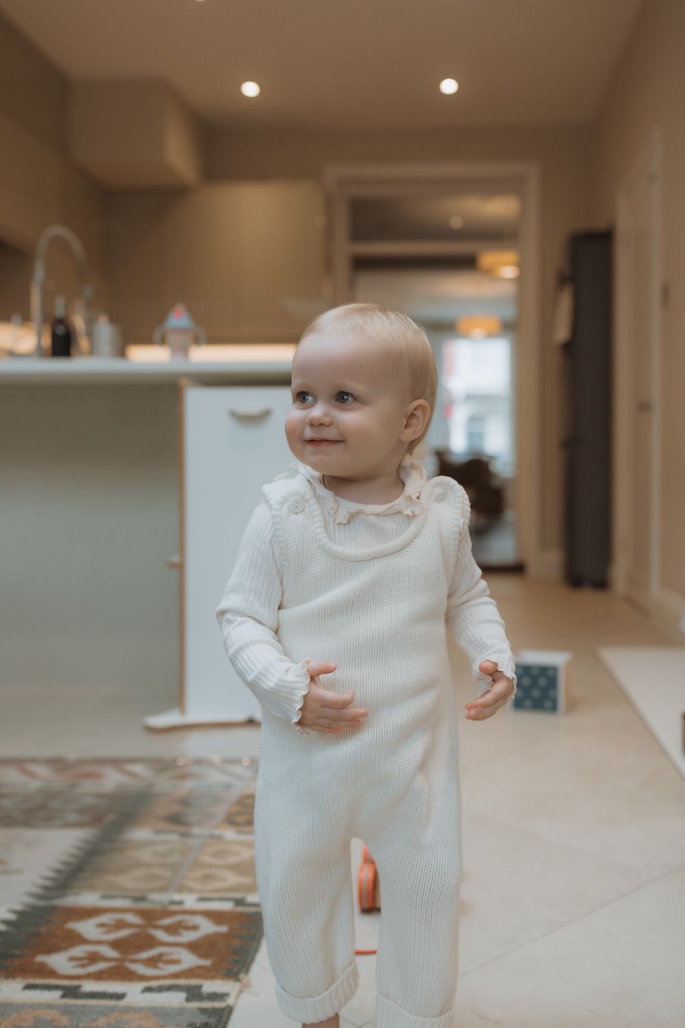 A small child wearing a white ruffle-collar romper smiles while standing on a patterned rug in a modern home interior.