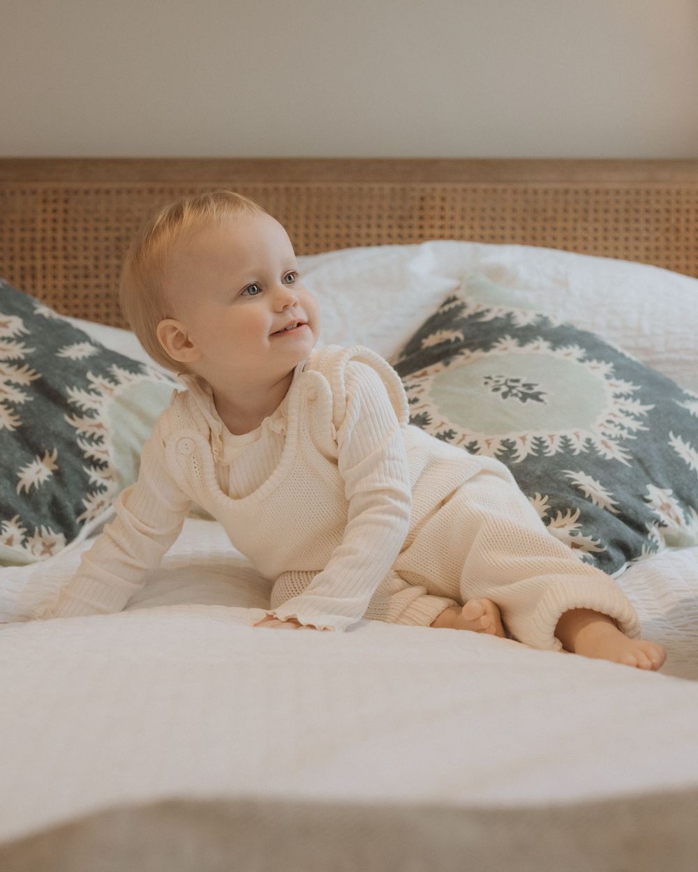 Baby in cream-colored outfit lying on white bedding with blue and green patterned pillows.
