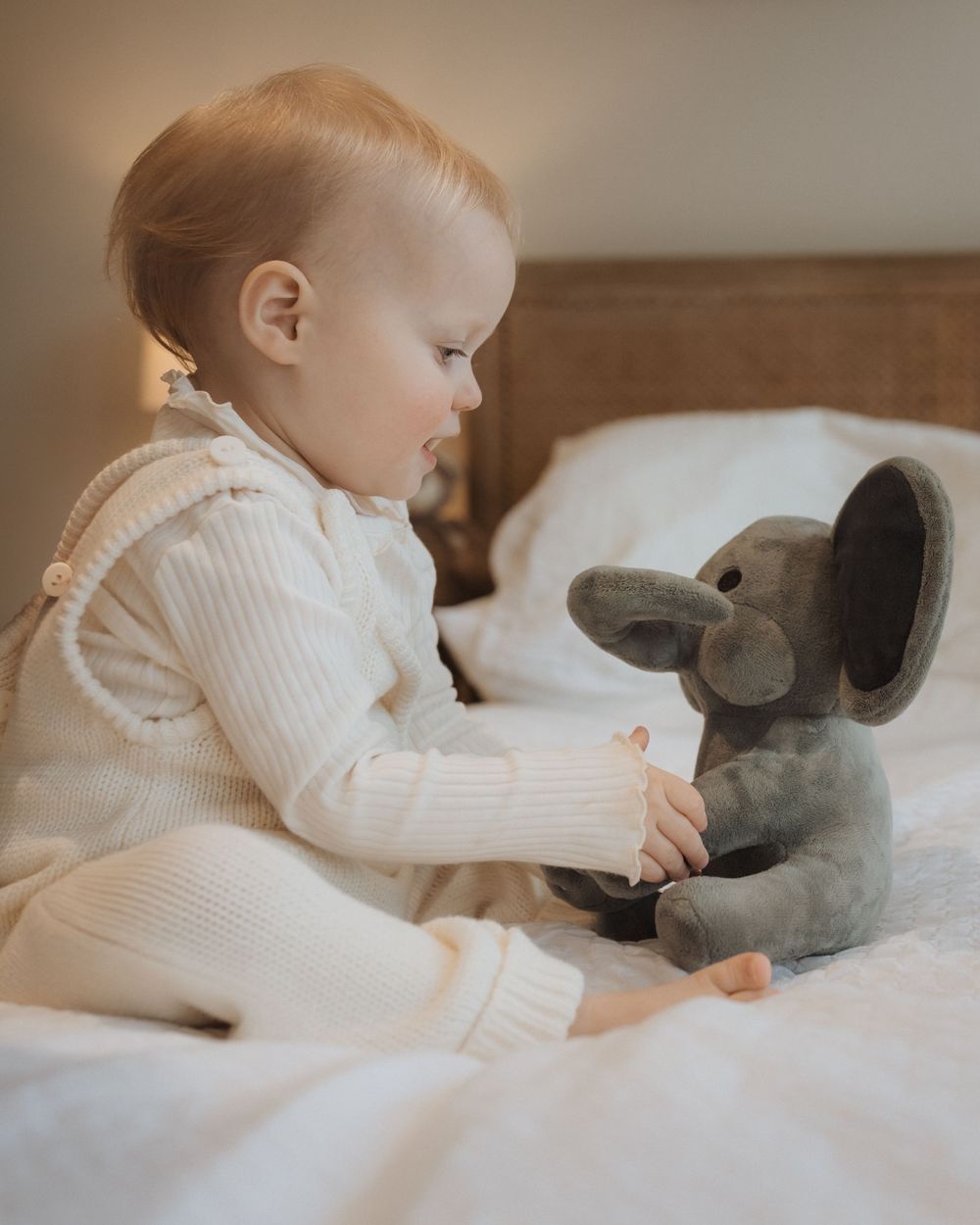 Baby wearing white knit outfit sits on bed looking at soft gray elephant stuffed animal toy.