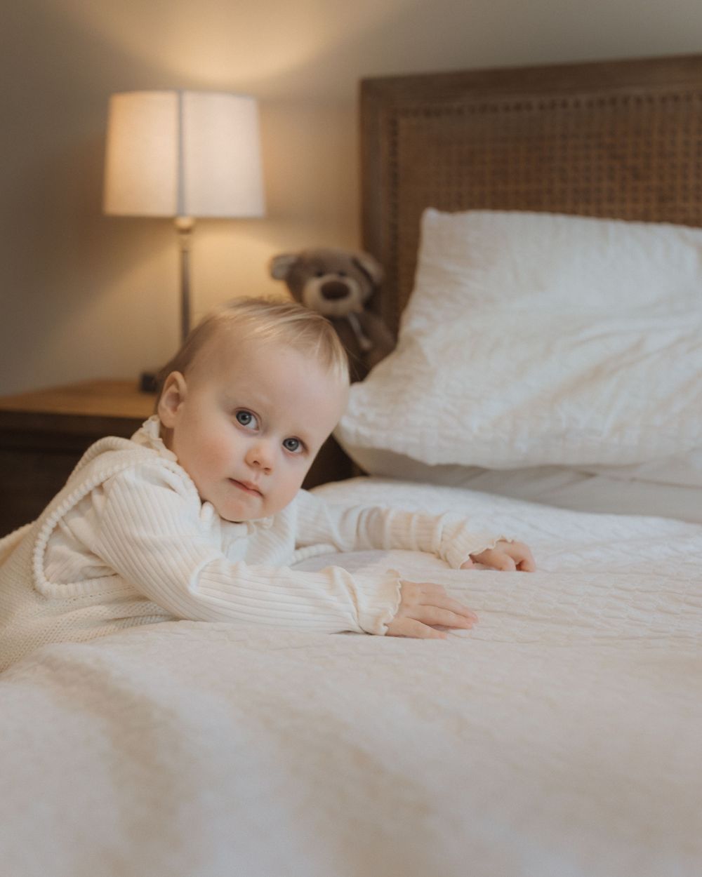 A small child in a white sweater rests on a cozy bed with white linens next to a warm bedside lamp.