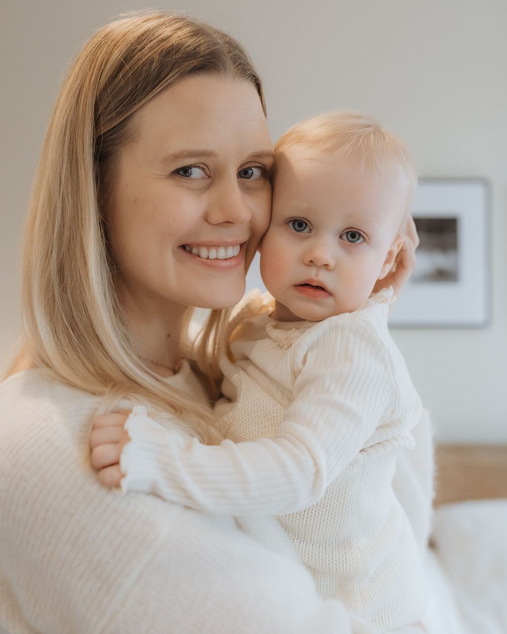 A parent wearing a cream-colored sweater holds a baby in a matching white sweater against a light background.