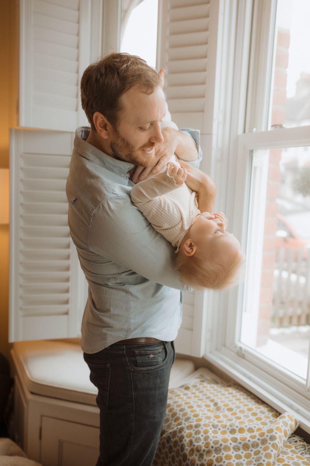 A series of tender moments between parent and infant in a sunlit room by a window with white shutters.
