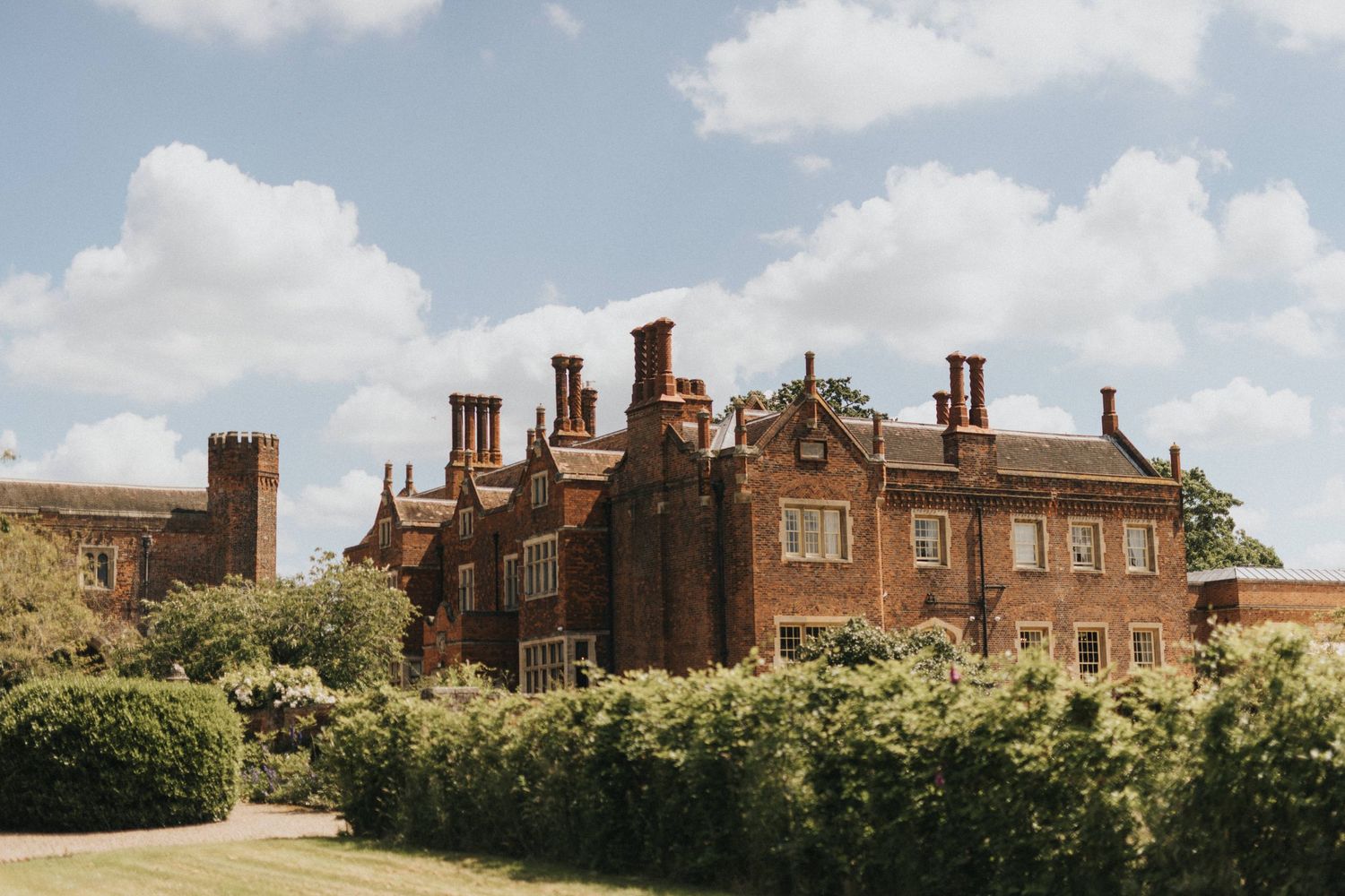 Historic red brick English manor house with ornate chimneys and Tudor architecture stands behind manicured hedges on a sunny day.