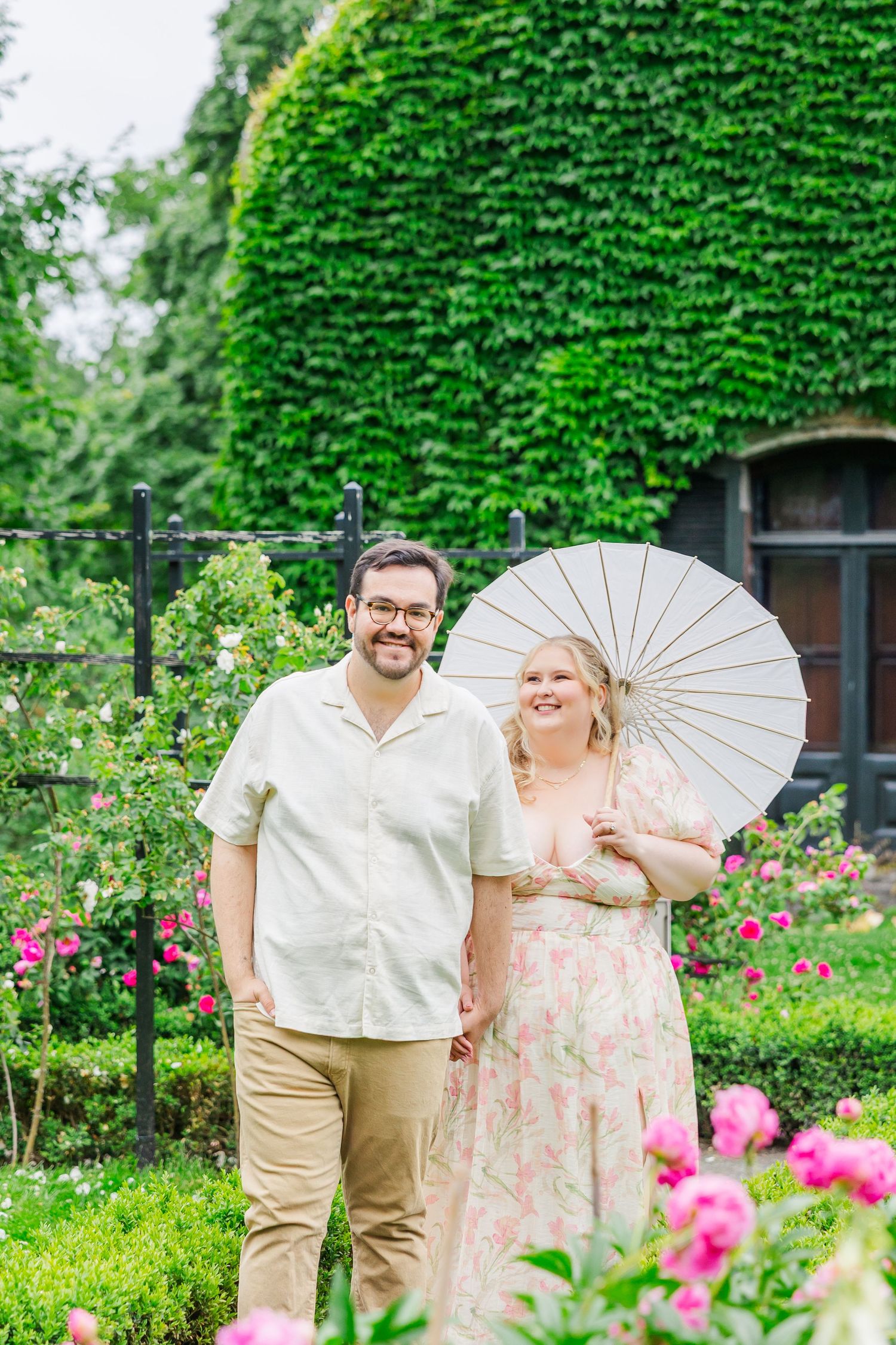 A couple stands in a vibrant garden with colorful pink flowers, while one holds a white parasol on a sunny day.