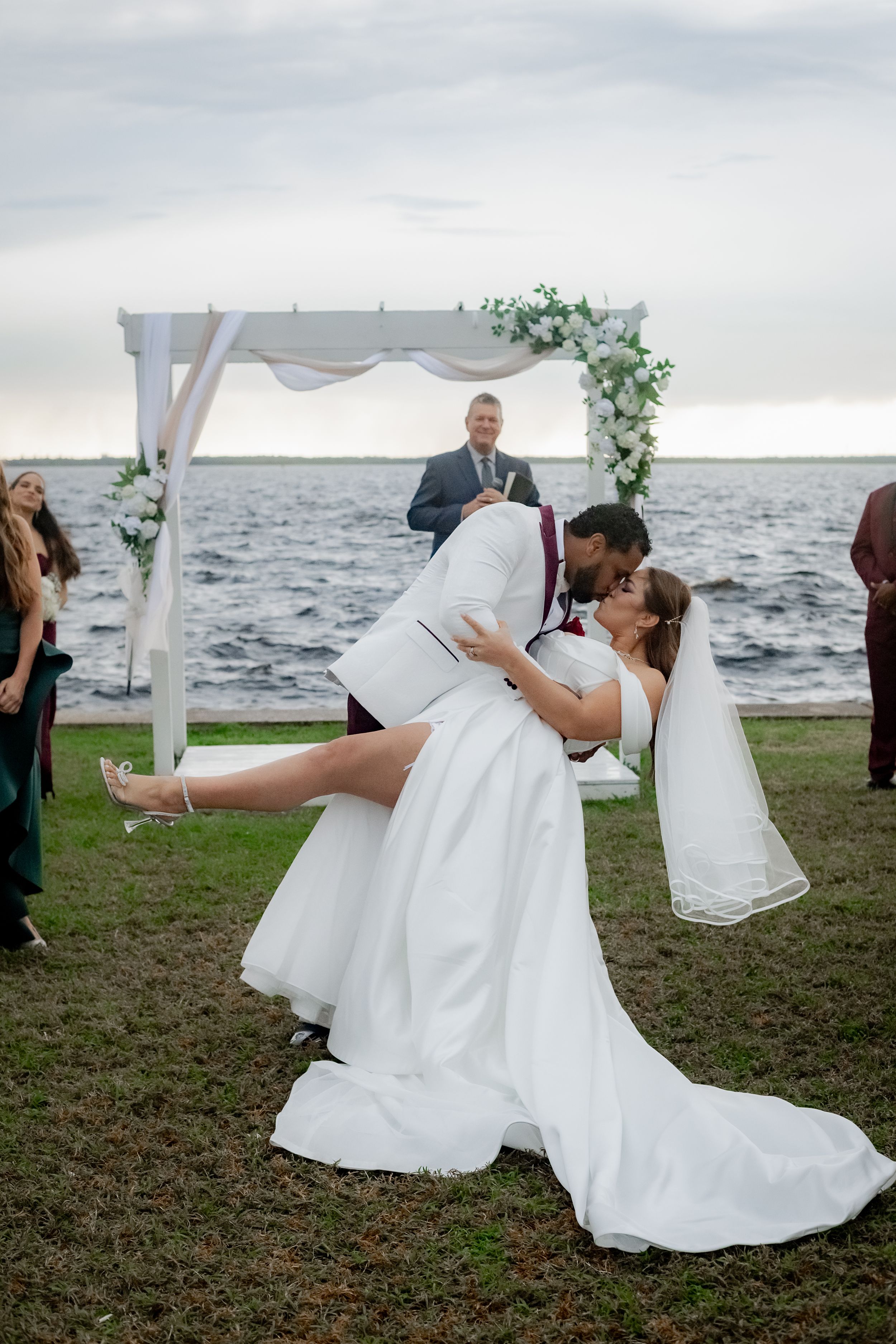 A romantic wedding ceremony takes place by the water under a white arch decorated with greenery at sunset.
