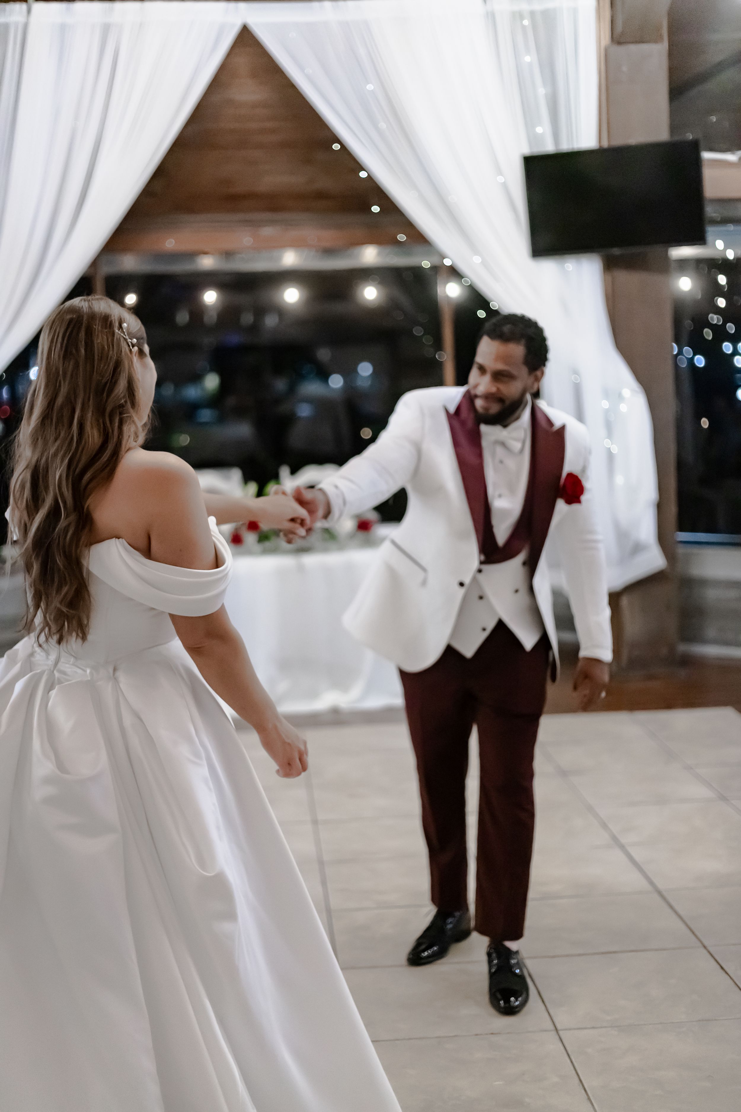 A couple in wedding attire dances together in a white-draped venue with romantic lighting.
