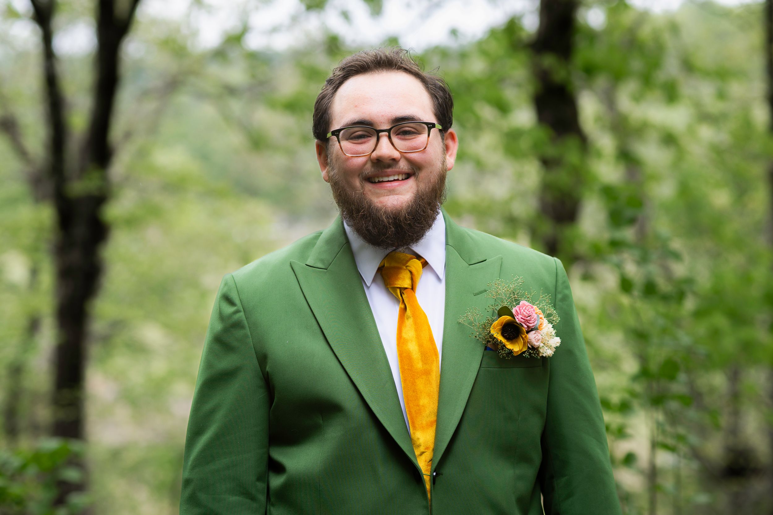 Groom in bright green suit jacket and yellow tie smiles outdoors in natural setting.