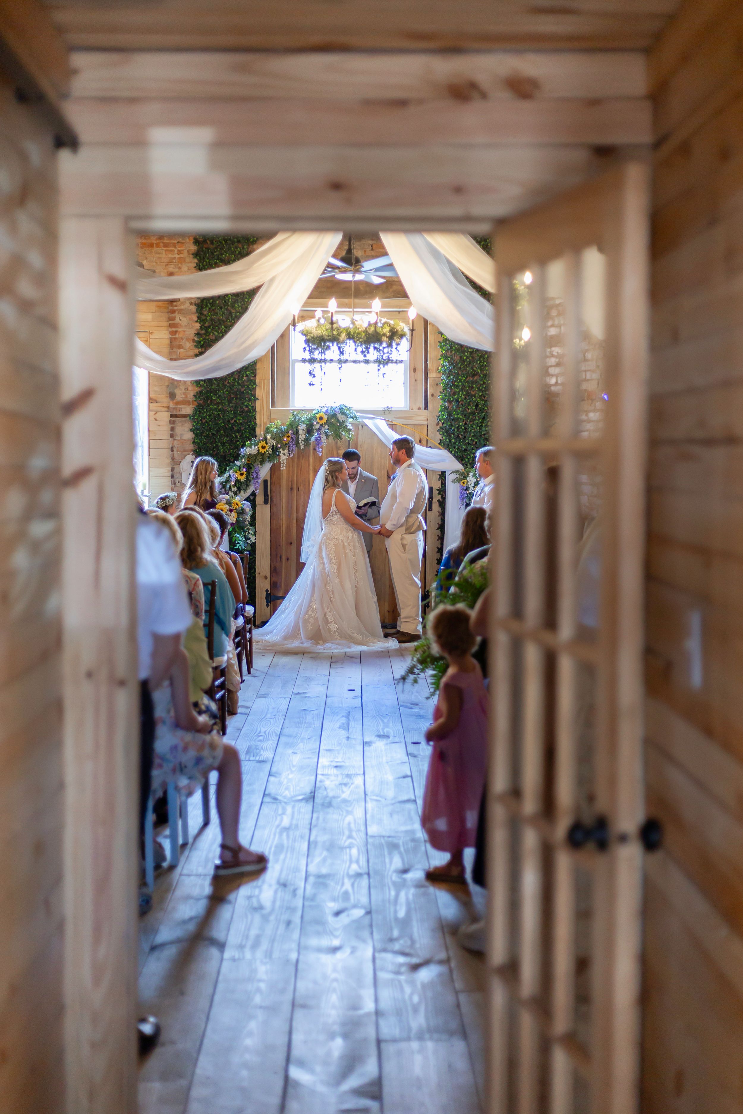 Wedding ceremony captured through wooden pillars in a rustic indoor venue.