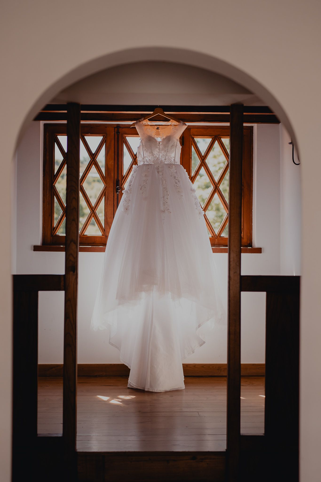 White wedding dress hanging in front of wooden arched windows in elegant venue.