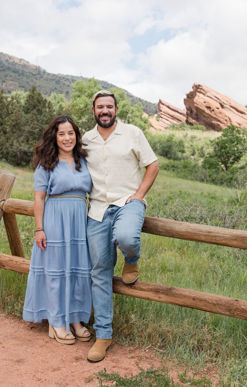 Couple stands by rustic wooden fence with scenic red rock formations and mountain landscape behind them.