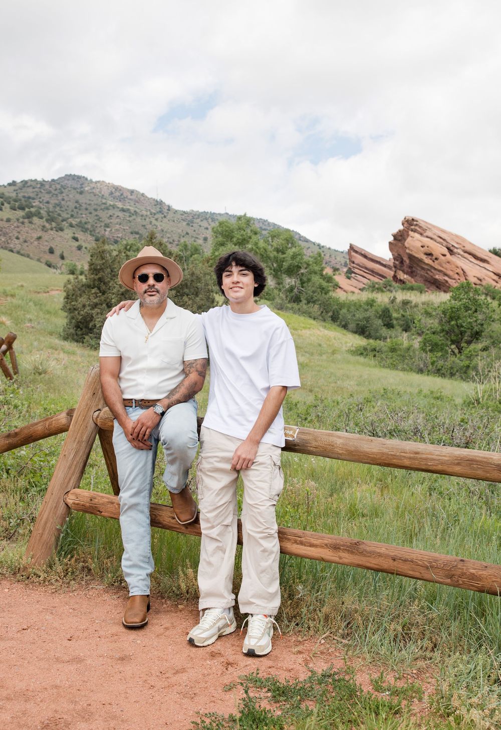 Two people sit together on a wooden fence with scenic Red Rocks formations and green hills in the background.