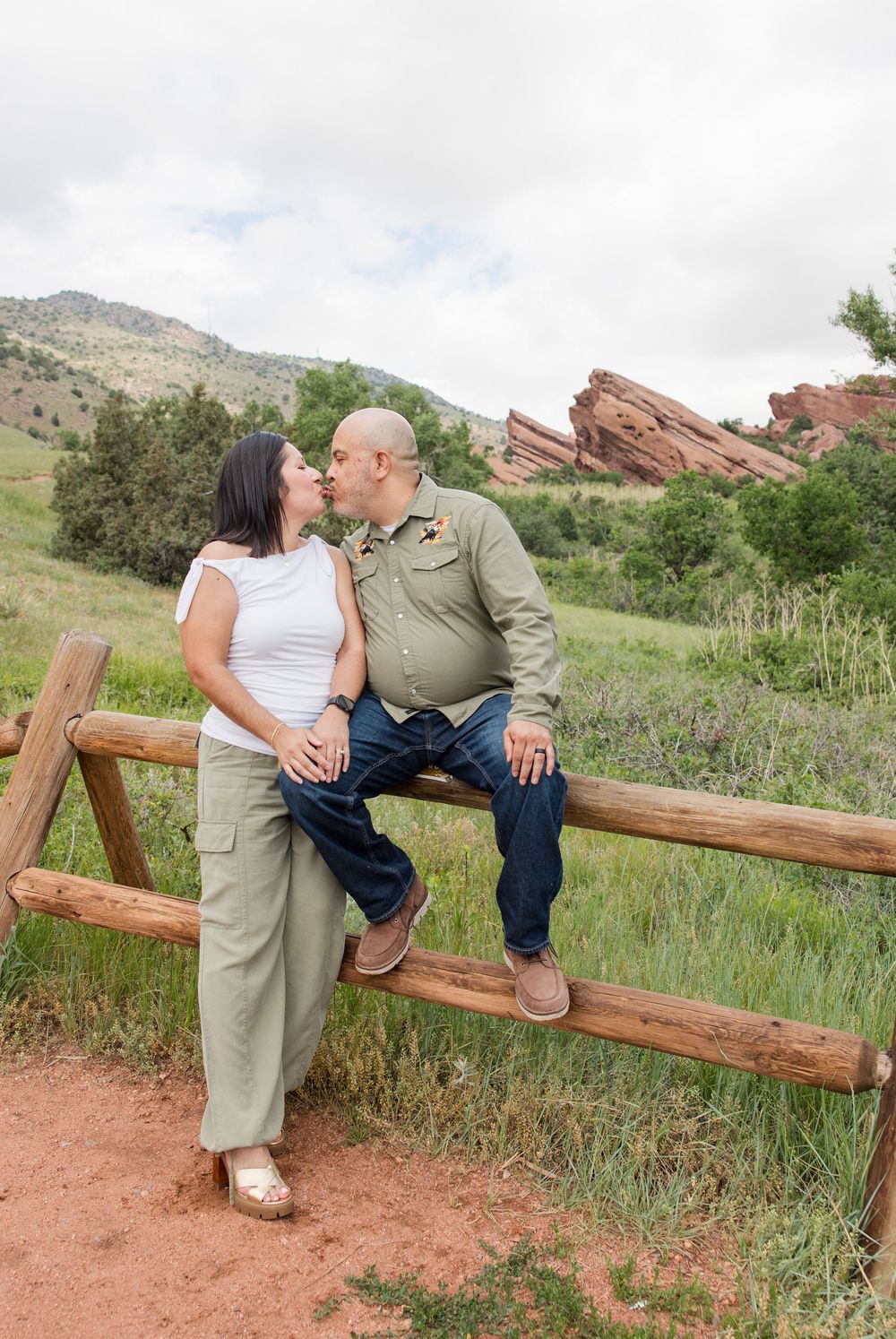 A couple sits together on a wooden fence at Red Rocks Park with mountains and red rock formations behind them.