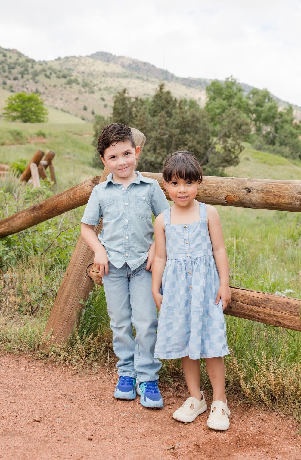 Young children in matching blue outfits sit and play by wooden fence at scenic mountain park.
