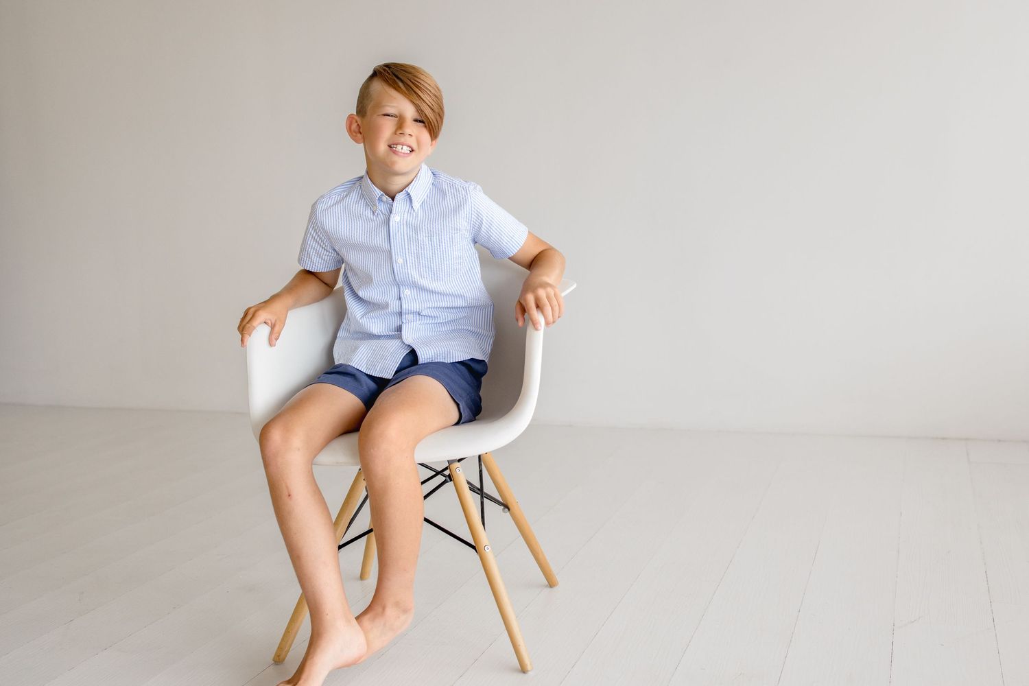 A casual portrait of someone sitting in a white chair wearing a light blue button-down shirt and shorts.