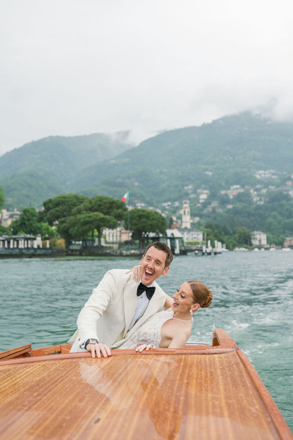 A couple relaxes on a classic wooden boat against a mountainous lake backdrop.