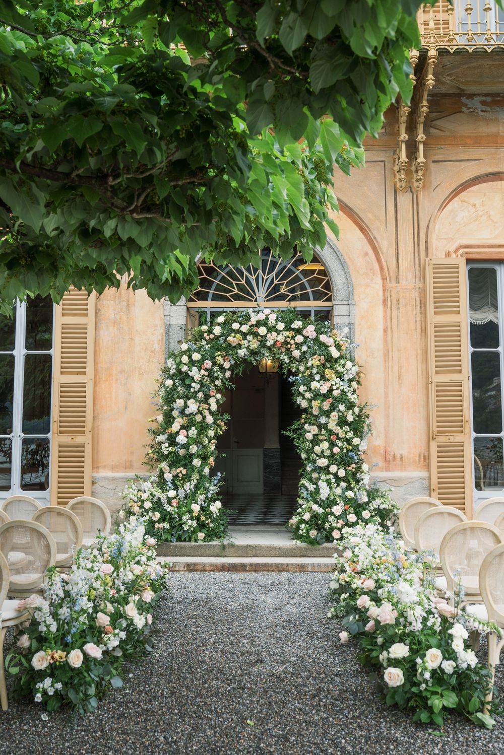White rose arches and elegant seating arrangements in a courtyard with cream-colored shuttered windows.