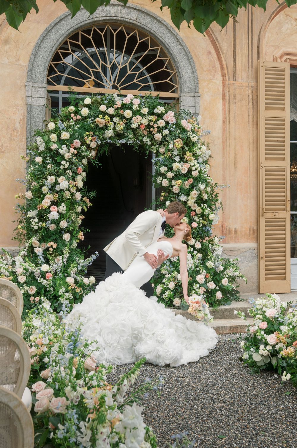 Wedding dress flowing on gravel courtyard beneath floral archways and historic architecture.