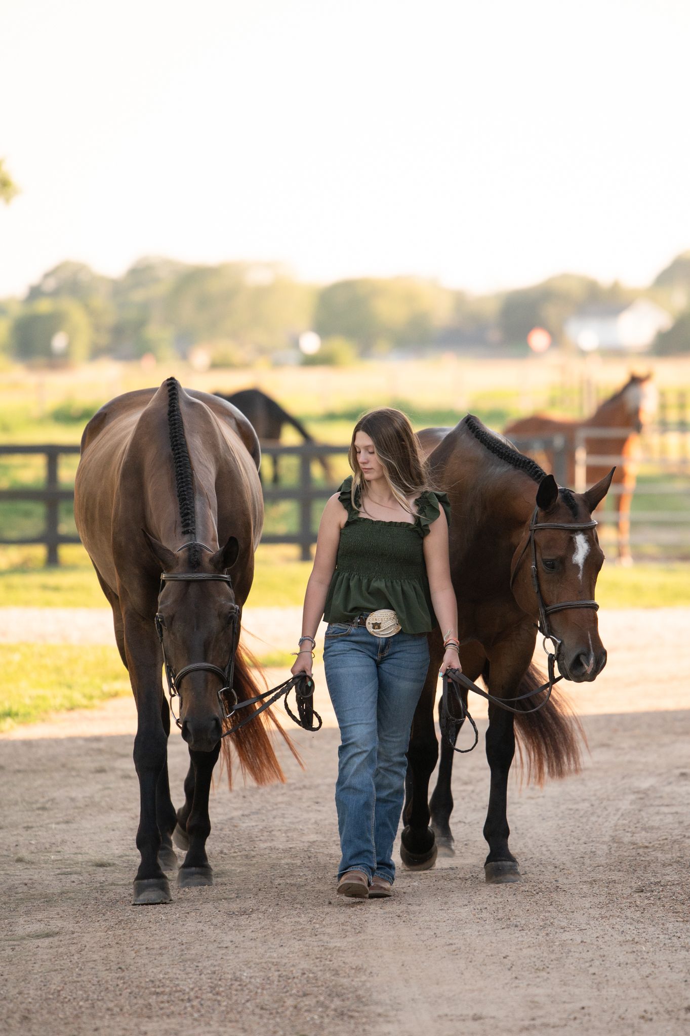 Woman in green top and jeans leads two dark horses along a dirt path at sunset on a peaceful farm.