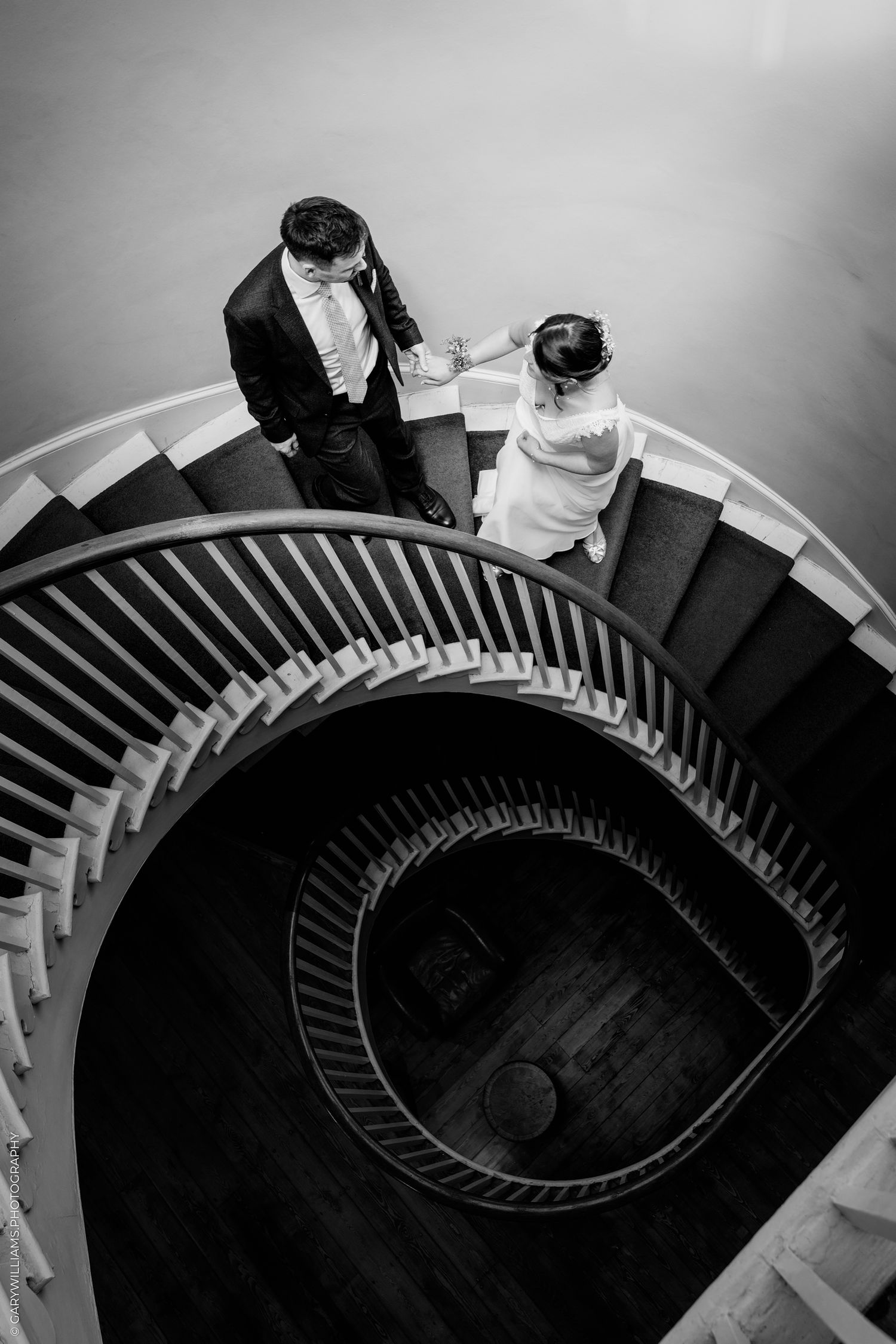 A black and white aerial view of a couple descending a dramatic spiral staircase in formal wedding attire.