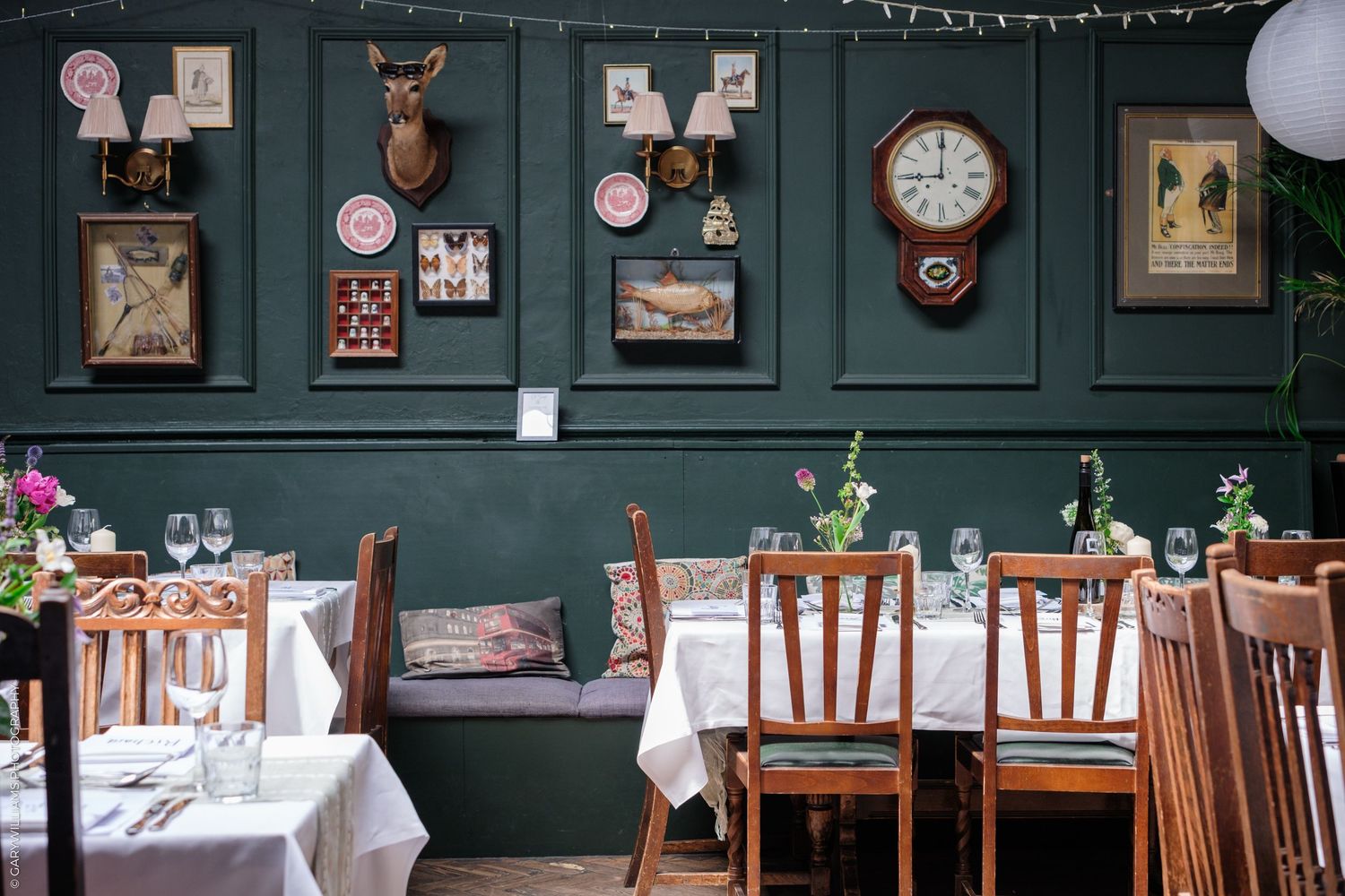 Cozy restaurant interior with dark green walls, wooden chairs, white tablecloths, and vintage decor including mounted clocks and artwork.