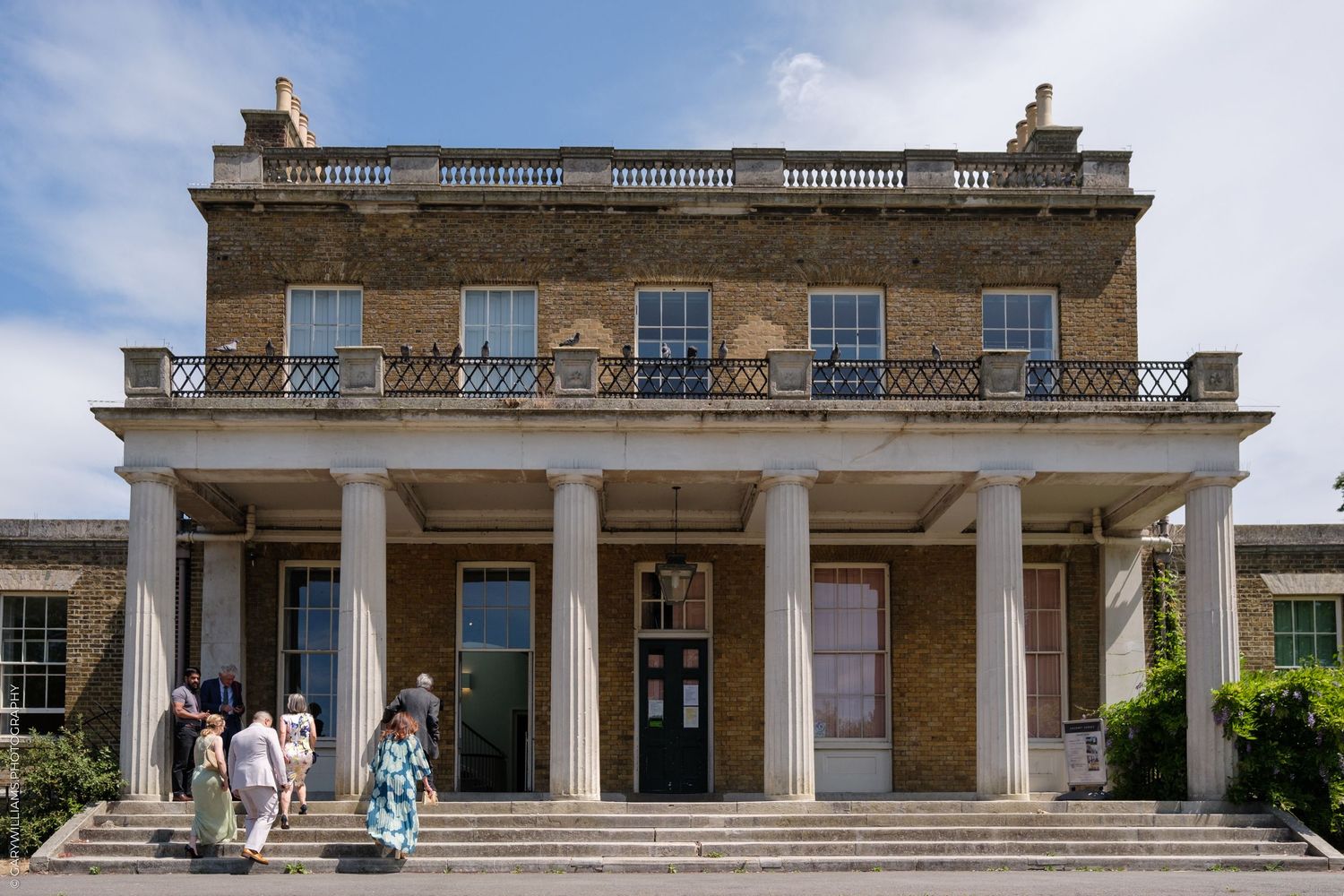 Elegant Georgian-style mansion with columned entrance portico and stone balustrade under bright blue sky.