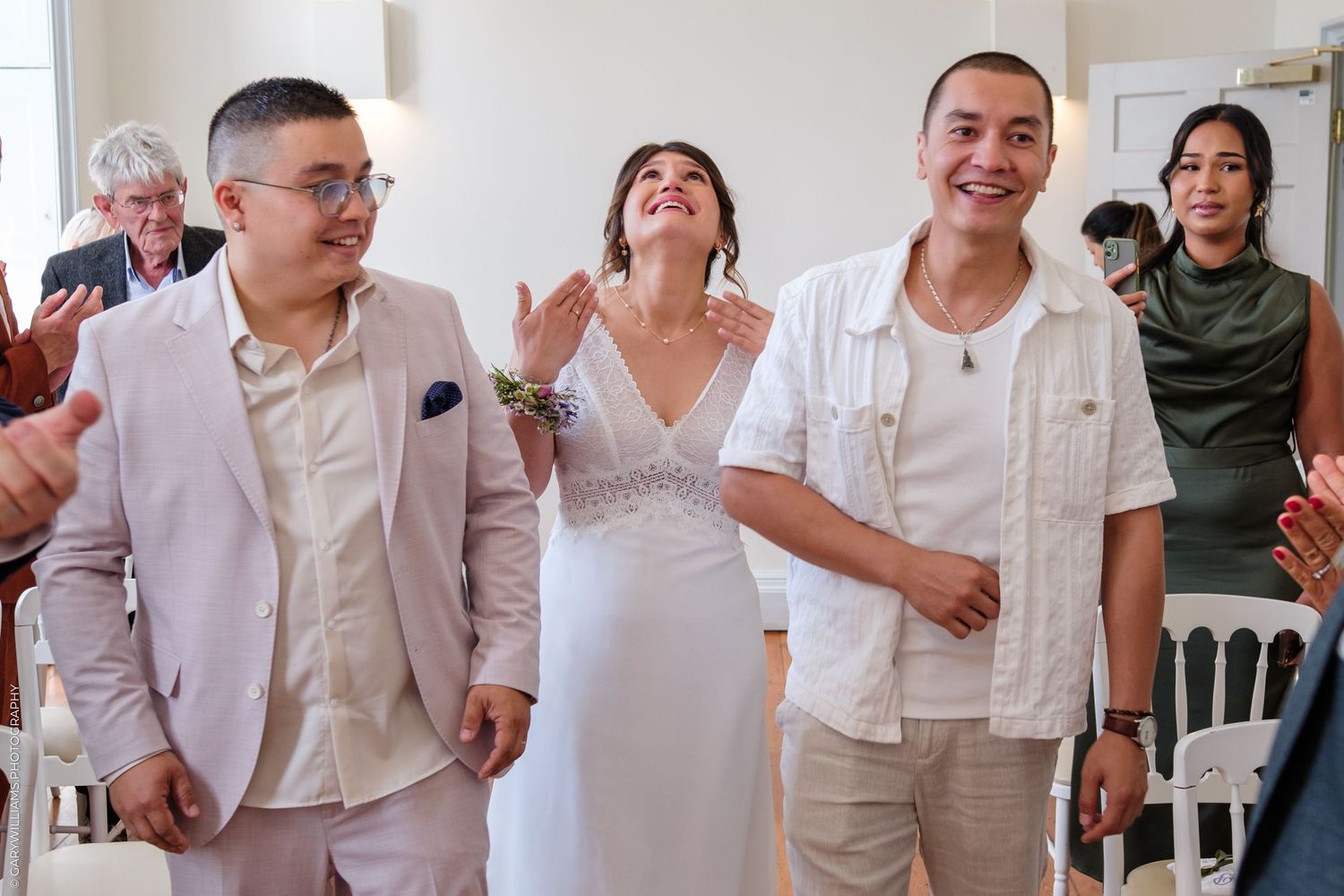 Joyful guests celebrate at a wedding reception wearing light colored outfits in an indoor venue.