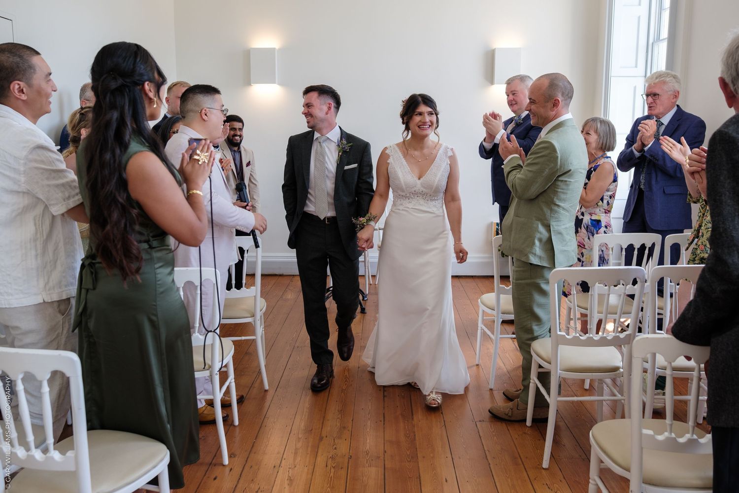 Wedding celebration in a bright indoor venue with guests lined up on white chairs as the couple walks down the aisle.