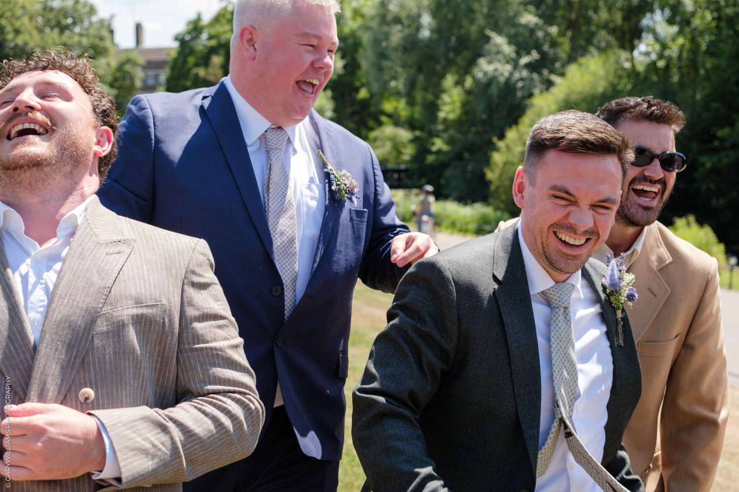 A group of wedding guests share a candid laugh together while standing outdoors on a sunny day.
