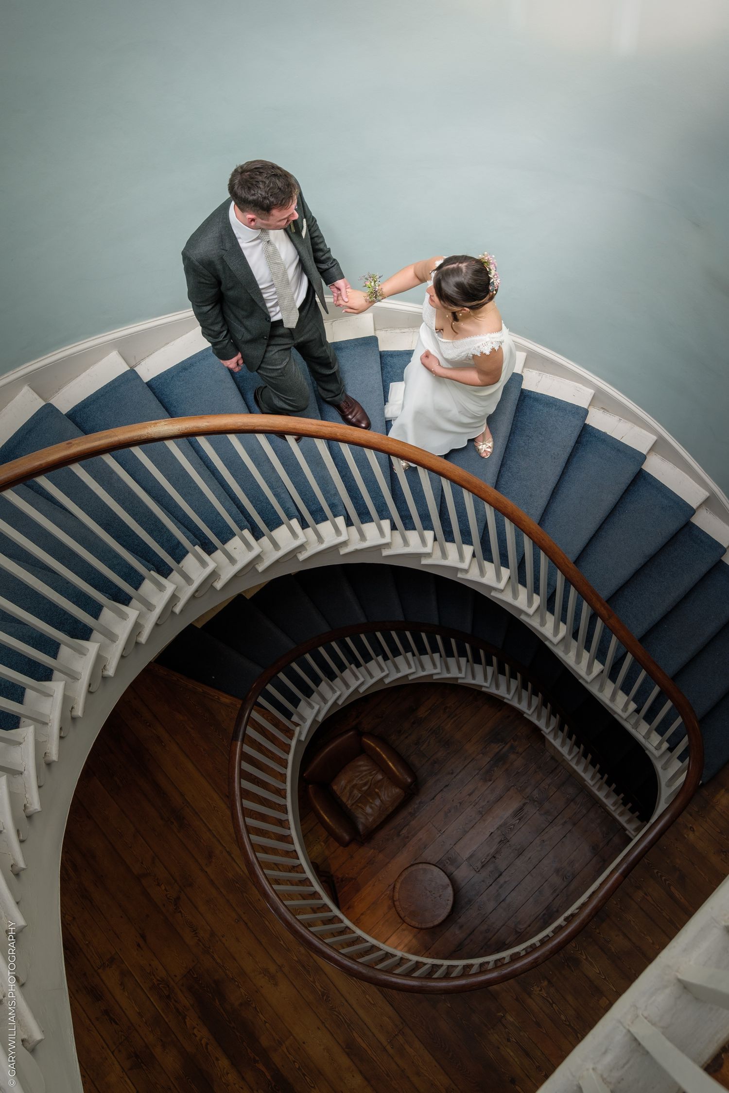 Aerial view of couple standing on elegant spiral staircase with navy blue carpet and dark wood floor.