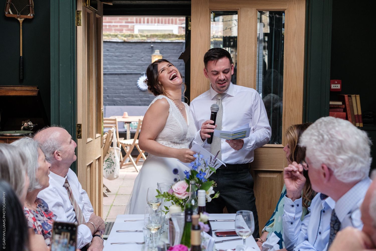 A couple shares a joyful moment while speaking at their wedding reception table surrounded by guests.