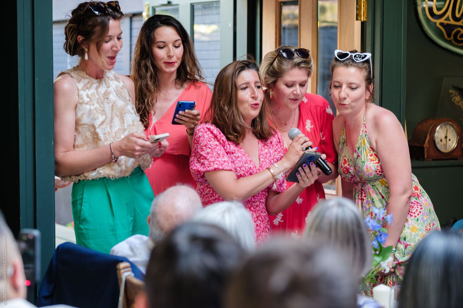 A group of women in colorful dresses and vintage-style clothing look at their phones together on the street.