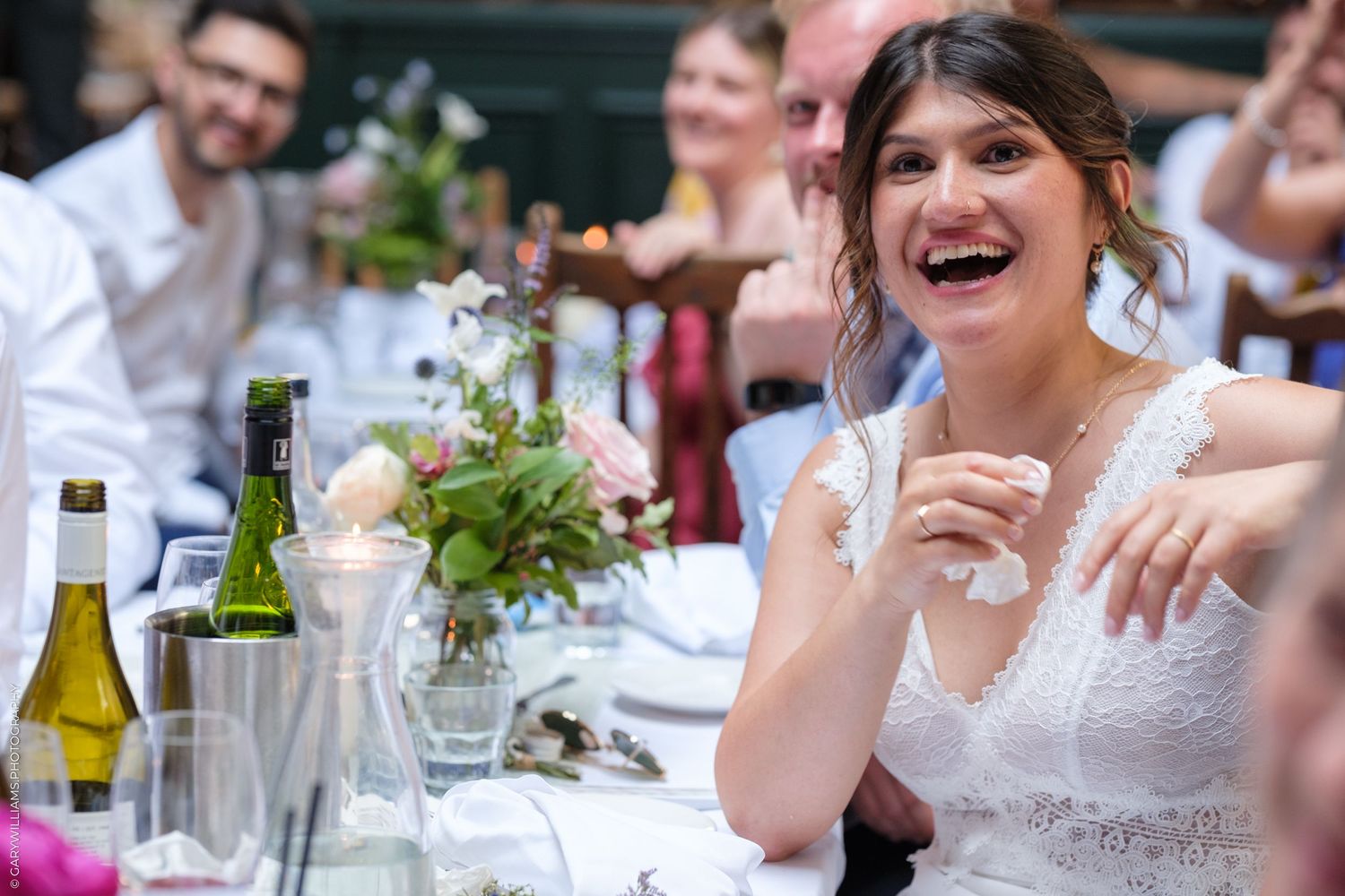 Wedding guests share laughter and conversation while dining at an outdoor reception table decorated with greenery.